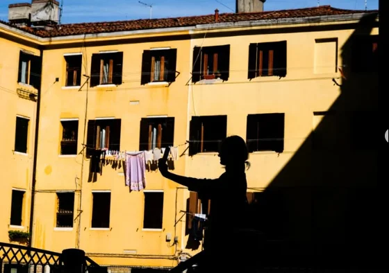Silhouette of a person taking a photo in front of a yellow apartment building with laundry hanging on a line, in deep shadow and strong sunlight. Photo by Manolis Soulos.