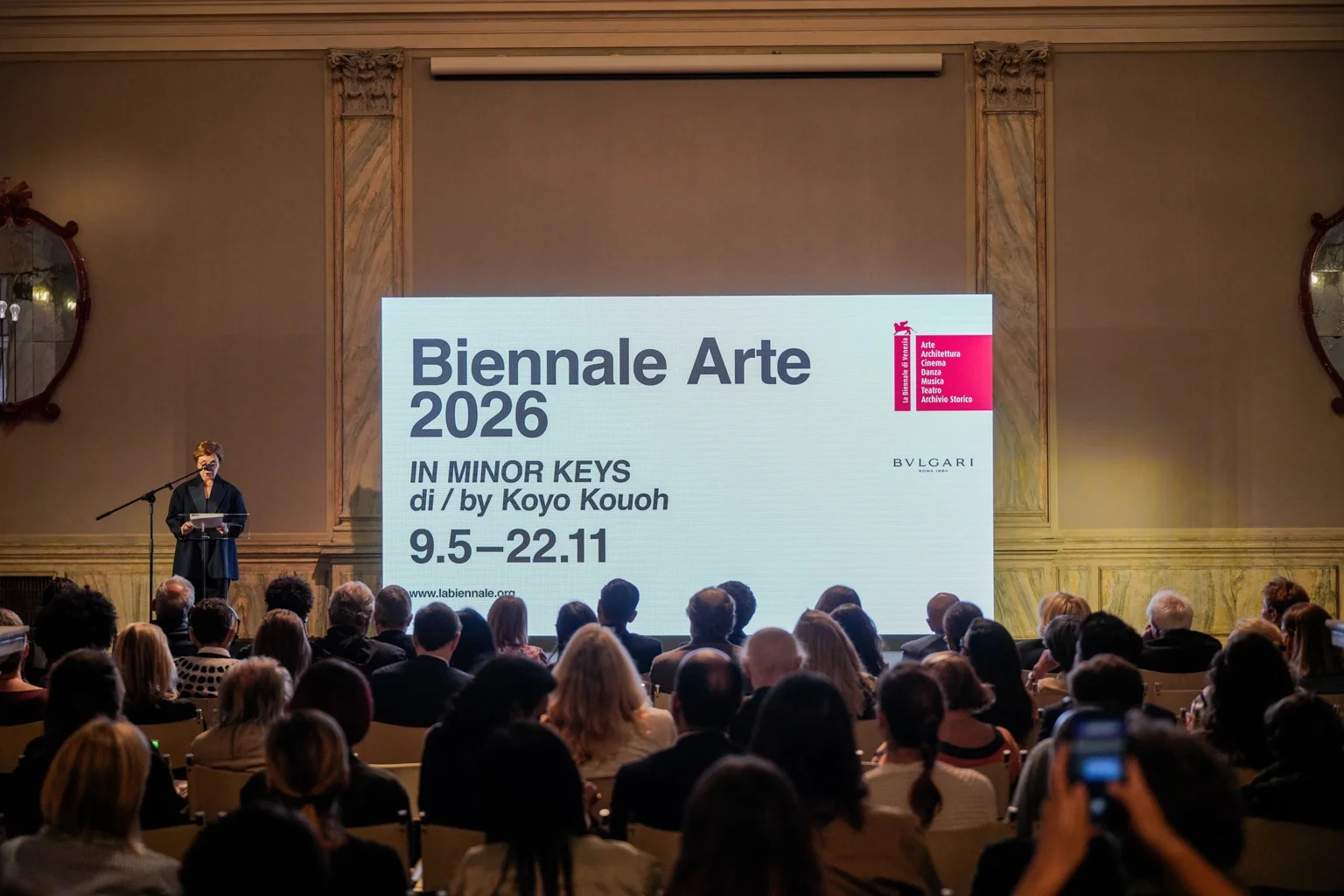 Presentation of Biennale Arte 2026 In Minor Keys in a grand hall, with a speaker at a podium and a large screen showing the exhibition dates in front of a seated audience. Photo by Andrea Avezz, courtesy of La Biennale di Venezia.