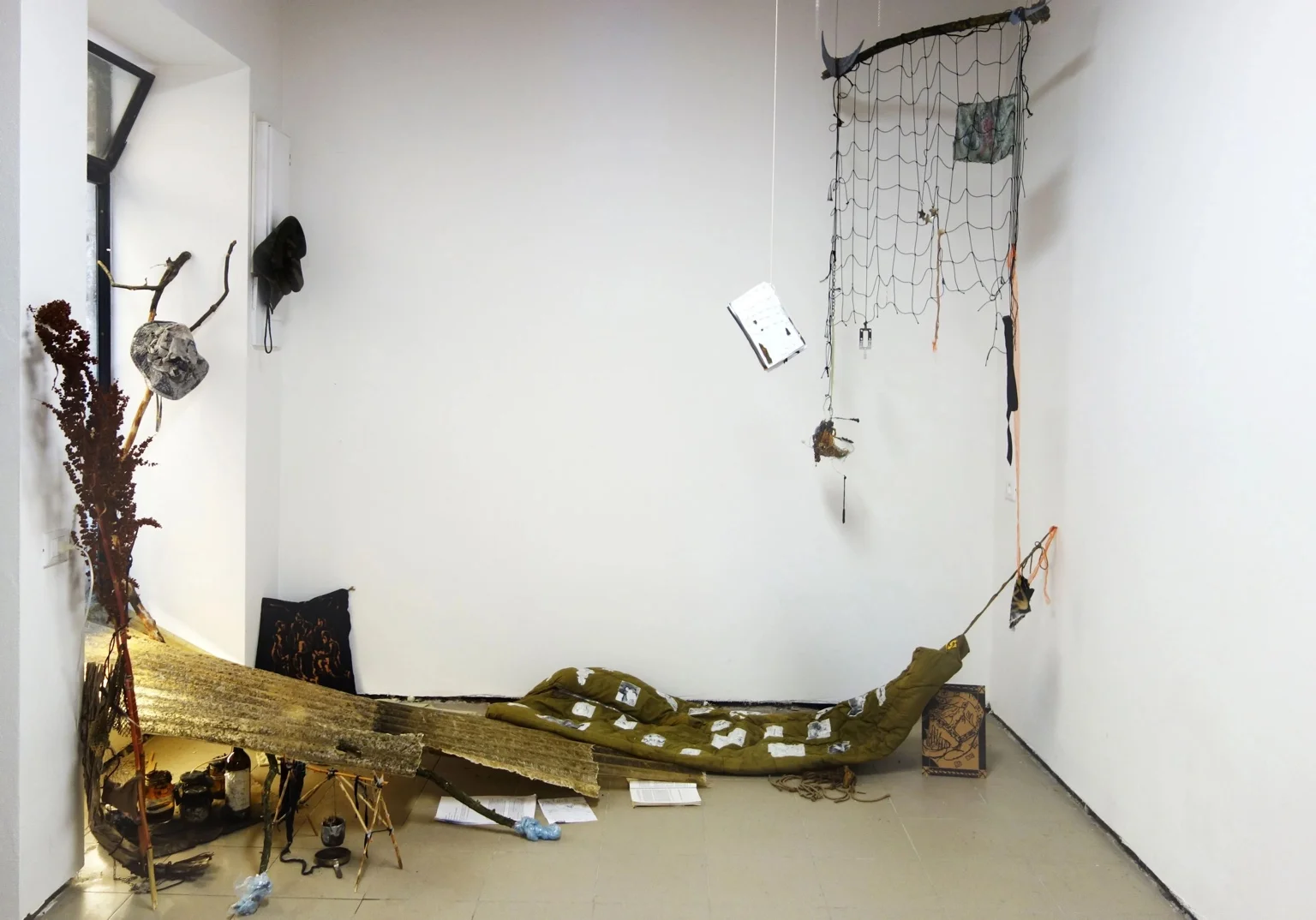 Installation view of Come raccogliere il fuoco che ci attraversa, with suspended nets, found objects, and a green padded form on the floor against a white gallery wall.