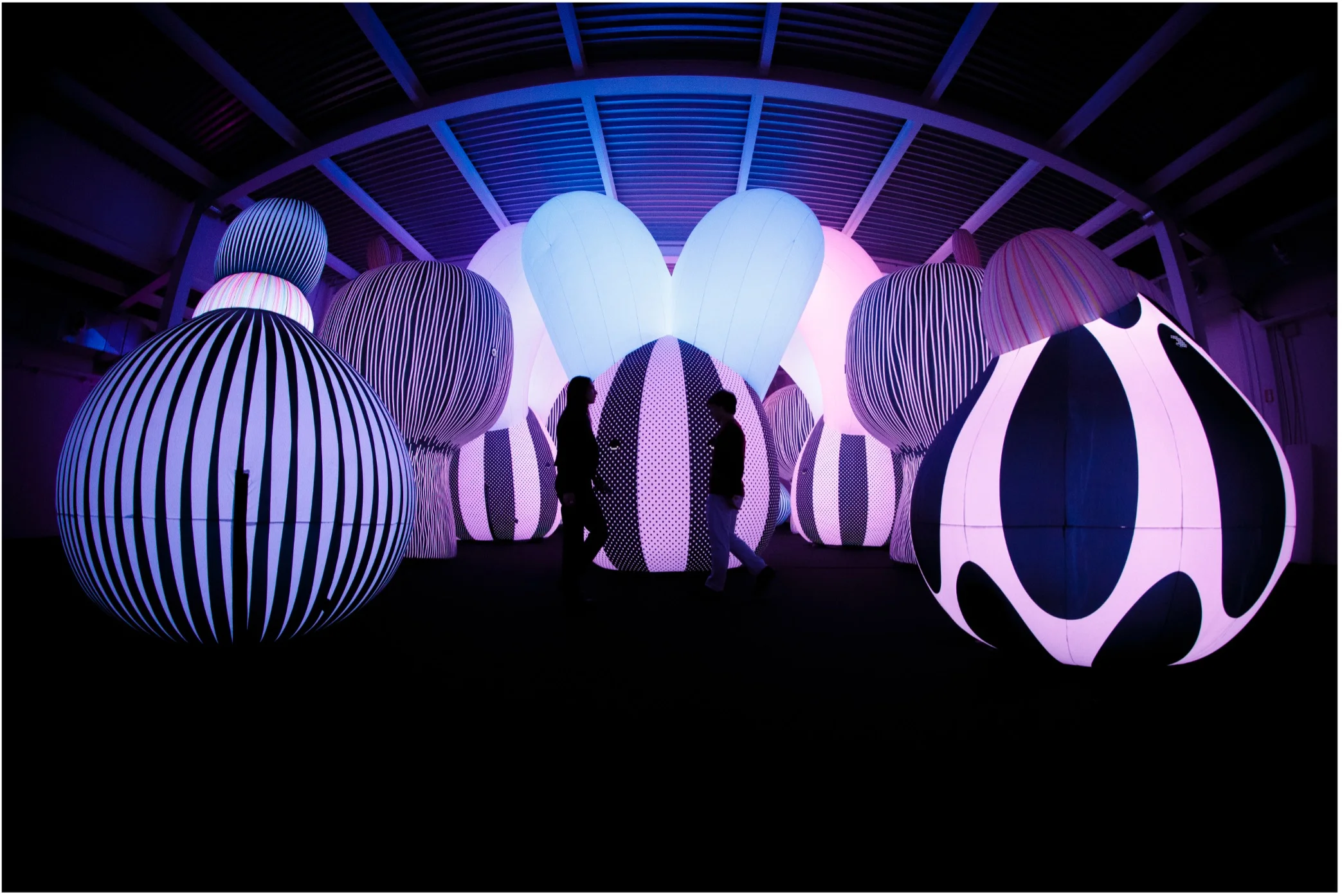 Immersive view of Airship Orchestra by ENESS at Balloon Museum, with visitors walking among glowing striped inflatable forms in purple and blue light.