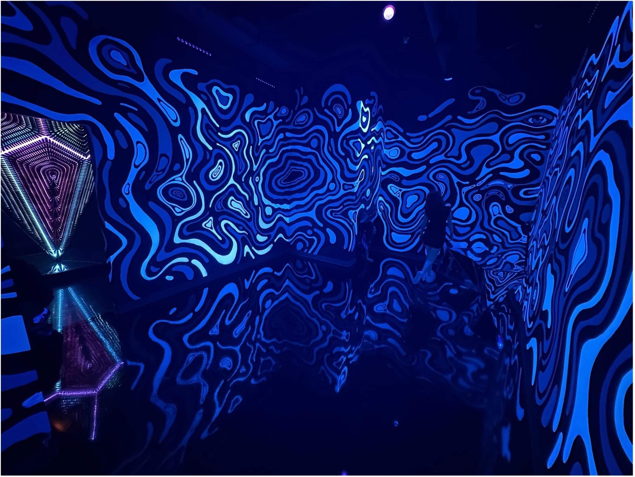Immersive dark room at Atlas 9 with swirling blue psychedelic wall patterns reflected on a glossy floor, and visitors walking through the glowing installation.