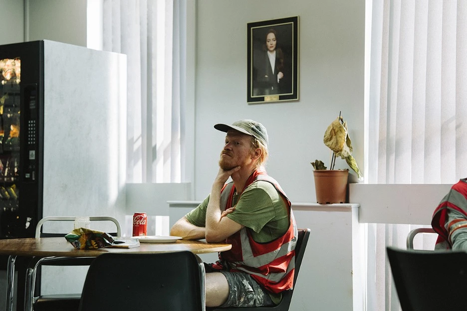 Jesse Plemons character in a high-visibility vest sits alone at a break-room table with snacks and a soda, staring thoughtfully ahead beneath a framed portrait on the wall.