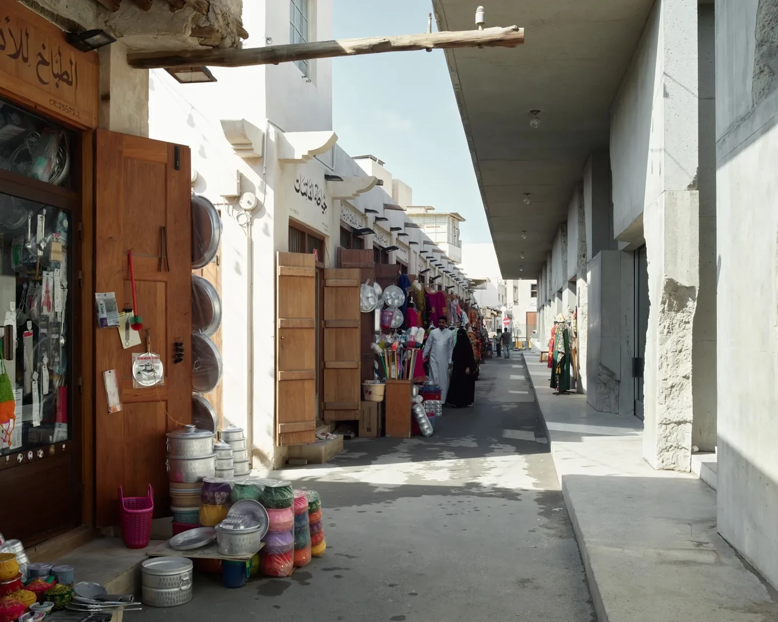 View along the Suq Al Qayssareyah and Ammarat in Muharraq, Bahrain, with market stalls, colourful household goods and clothing spilling into a narrow alleyway, framed by traditional wooden doors on one side and the rough concrete facade by Studio Anne Holtrop on the other.