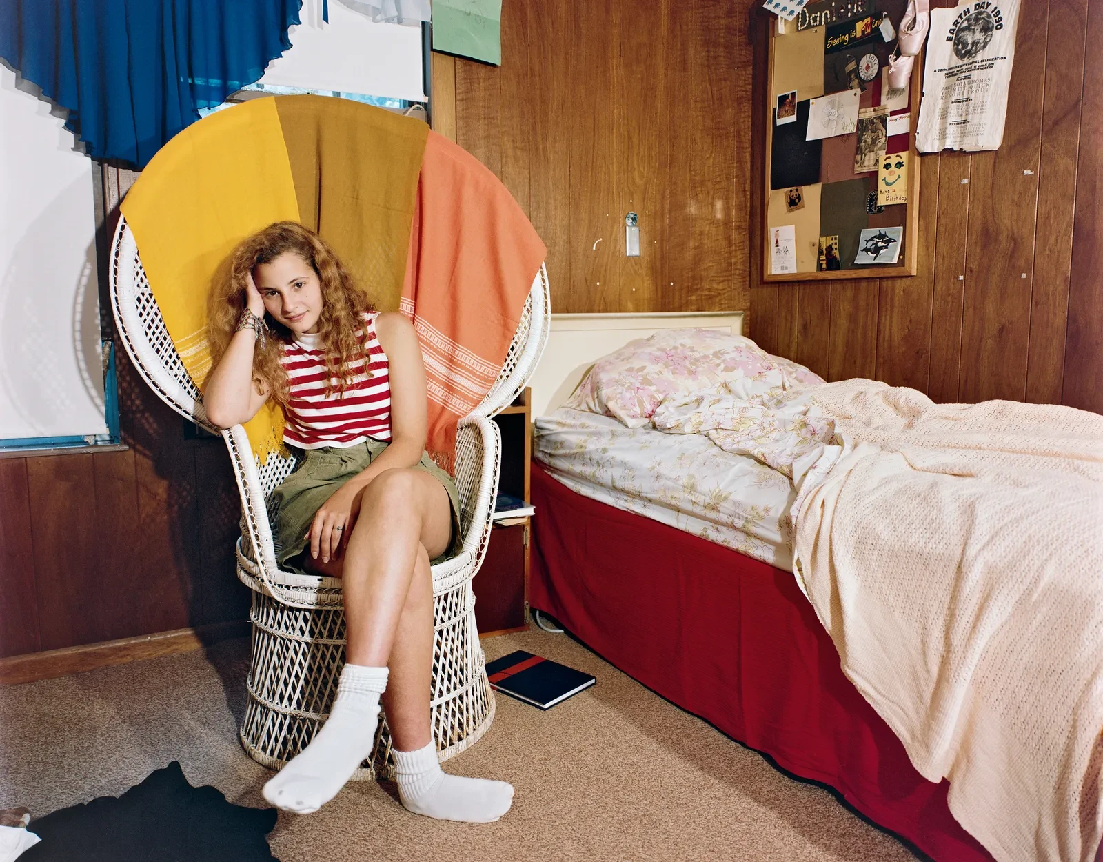 Teenage girl sitting in a wicker chair beside her bed in a wood-paneled bedroom, photographed by Adrienne Salinger in the 1990s.