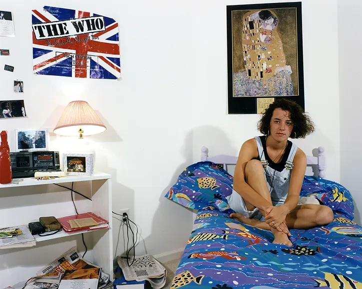 Teenage girl sitting cross-legged on her bed in a brightly decorated bedroom with music posters, photographed by Adrienne Salinger in the 1990s.