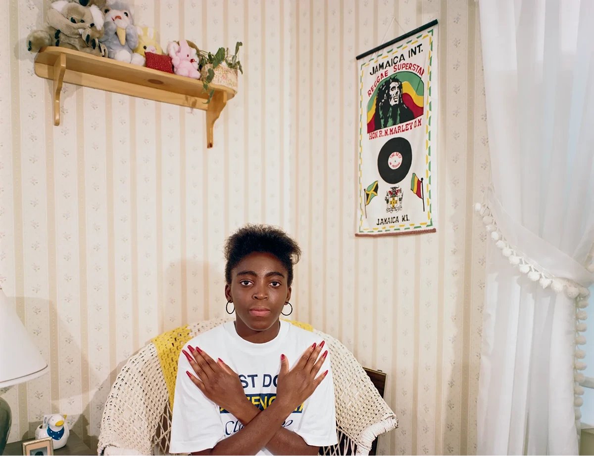 Teenage girl sitting in a wicker chair in her bedroom with arms crossed, photographed by Adrienne Salinger for the Teenagers in Their Bedrooms series.