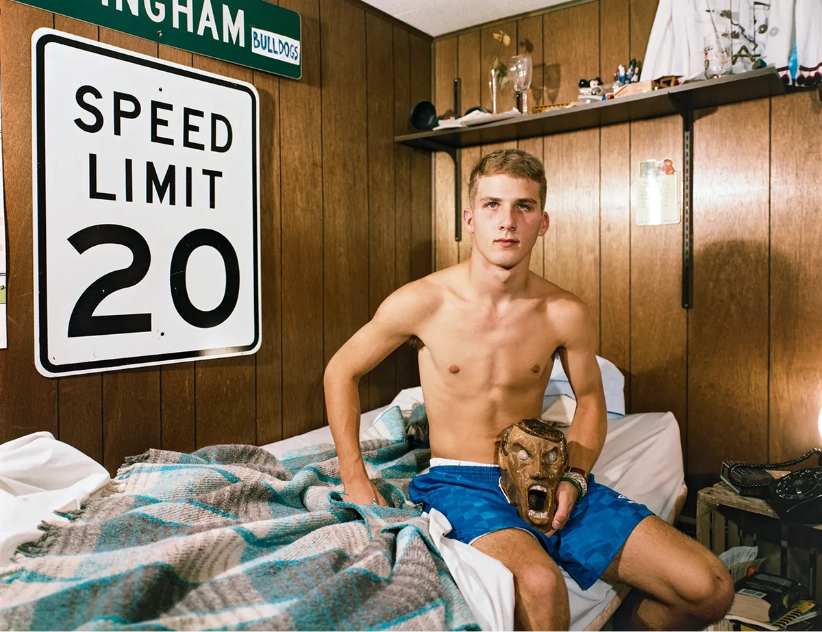 Teenage boy sitting on his bed holding a carved mask in a wood-paneled bedroom, photographed by Adrienne Salinger in the 1990s.