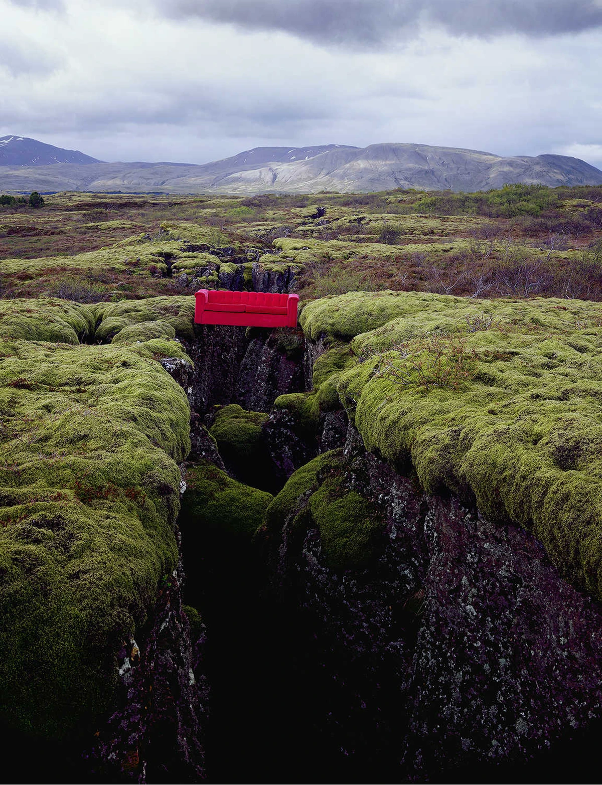 Red couch placed across a rocky crevice in a moss-covered landscape, photographed by Horst Wackerbarth for The Red Couch project.