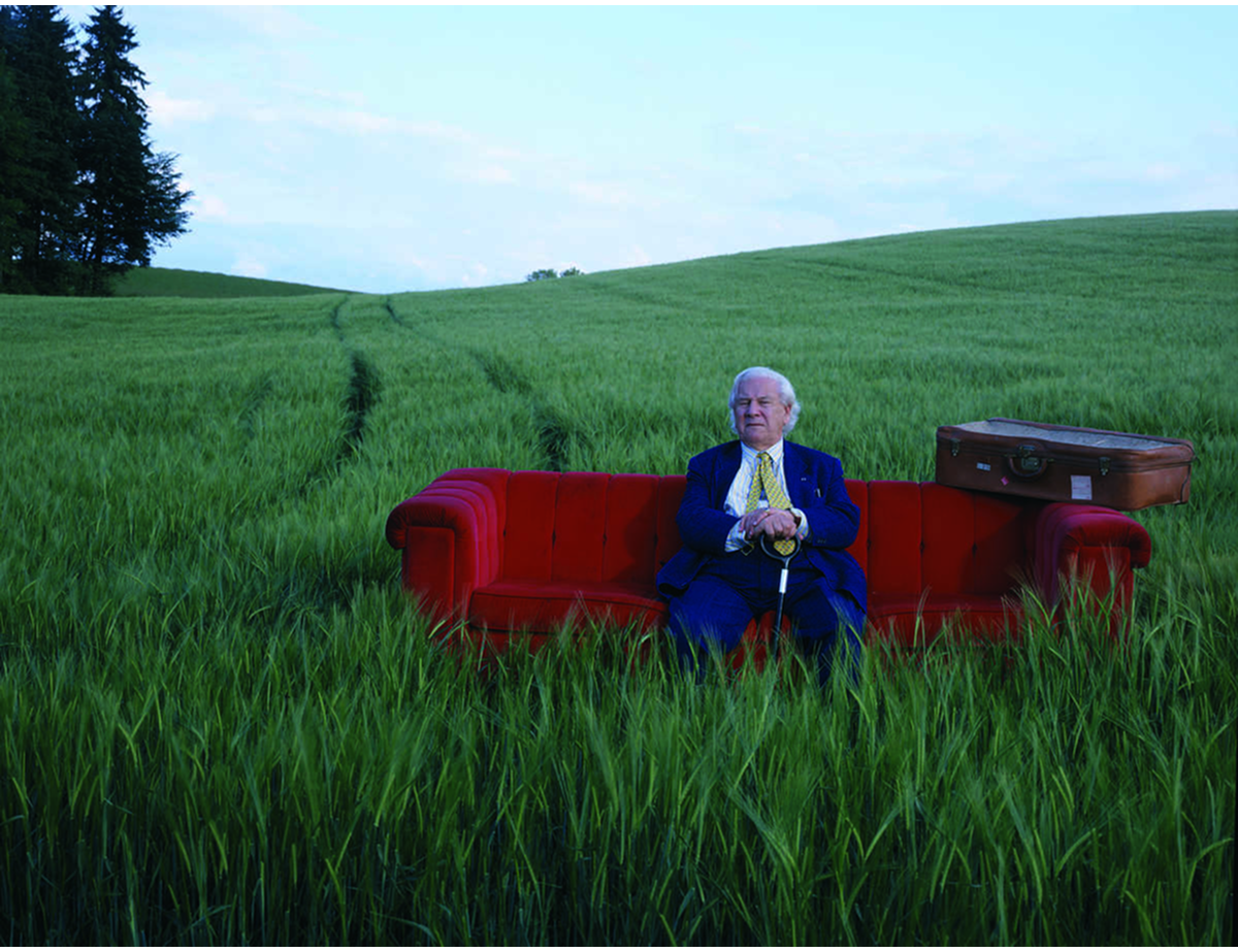 Sir Peter Ustinov sitting on a red couch in a green field, photographed by Horst Wackerbarth for The Red Couch project.