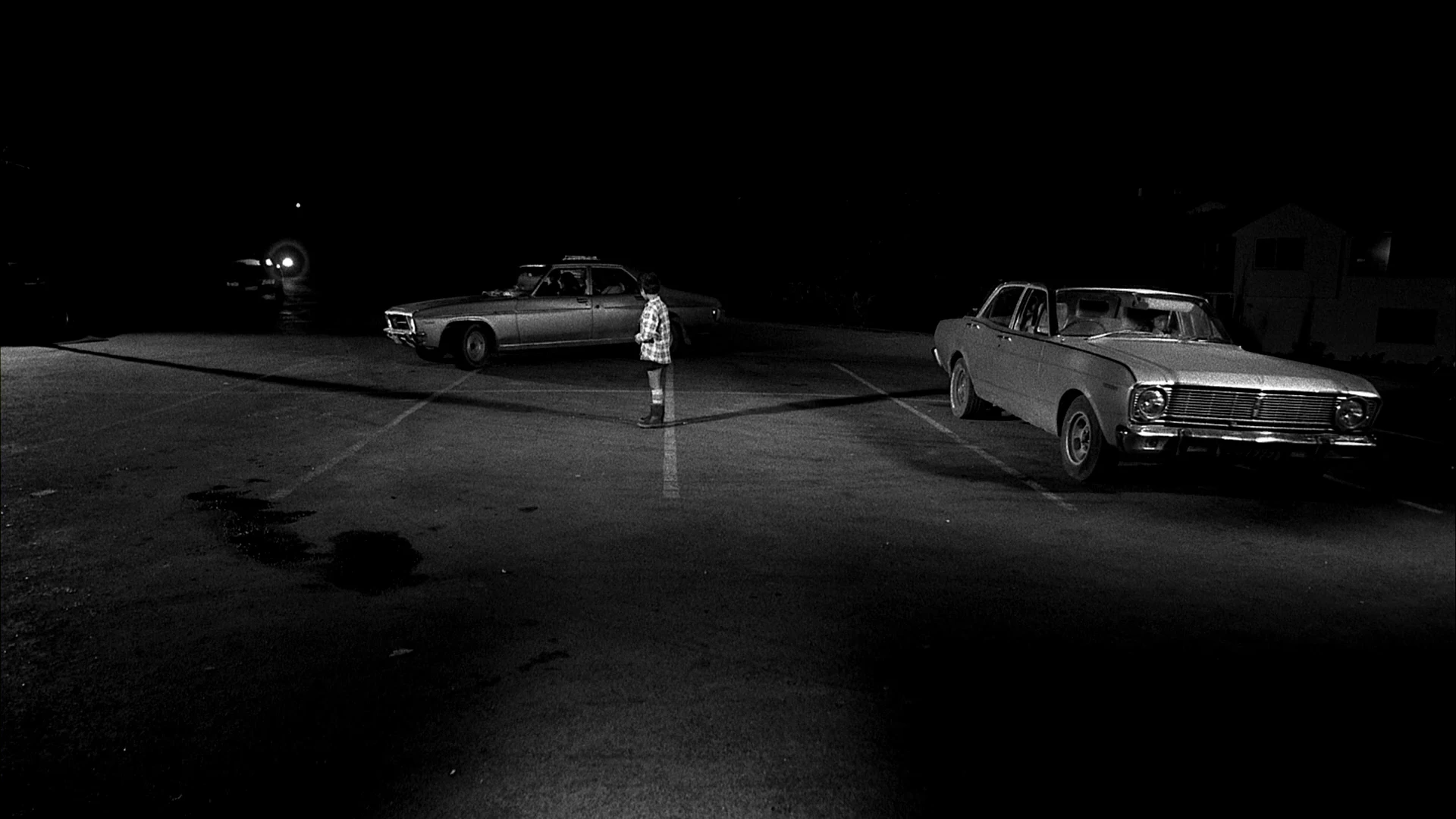 Black-and-white still from Two Cars, One Night (2003) showing a child standing between two parked cars at night, emphasizing quiet intimacy and cinematic minimalism.