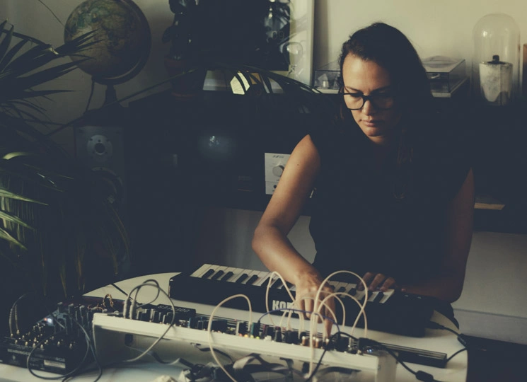 Christina Vantzou working at a home studio desk in 2018, seated at a keyboard and modular equipment with patch cables, surrounded by plants and audio gear in soft natural light.