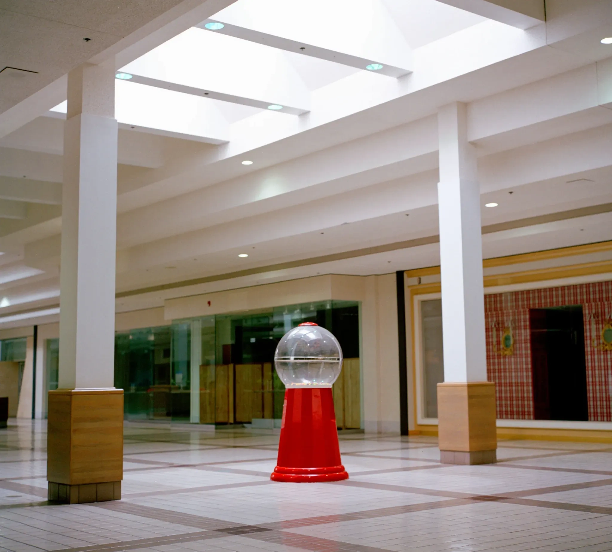 Interior of an abandoned shopping mall with tiled floors, white columns and skylights, centered on a red gumball machine standing alone in an otherwise empty retail space.