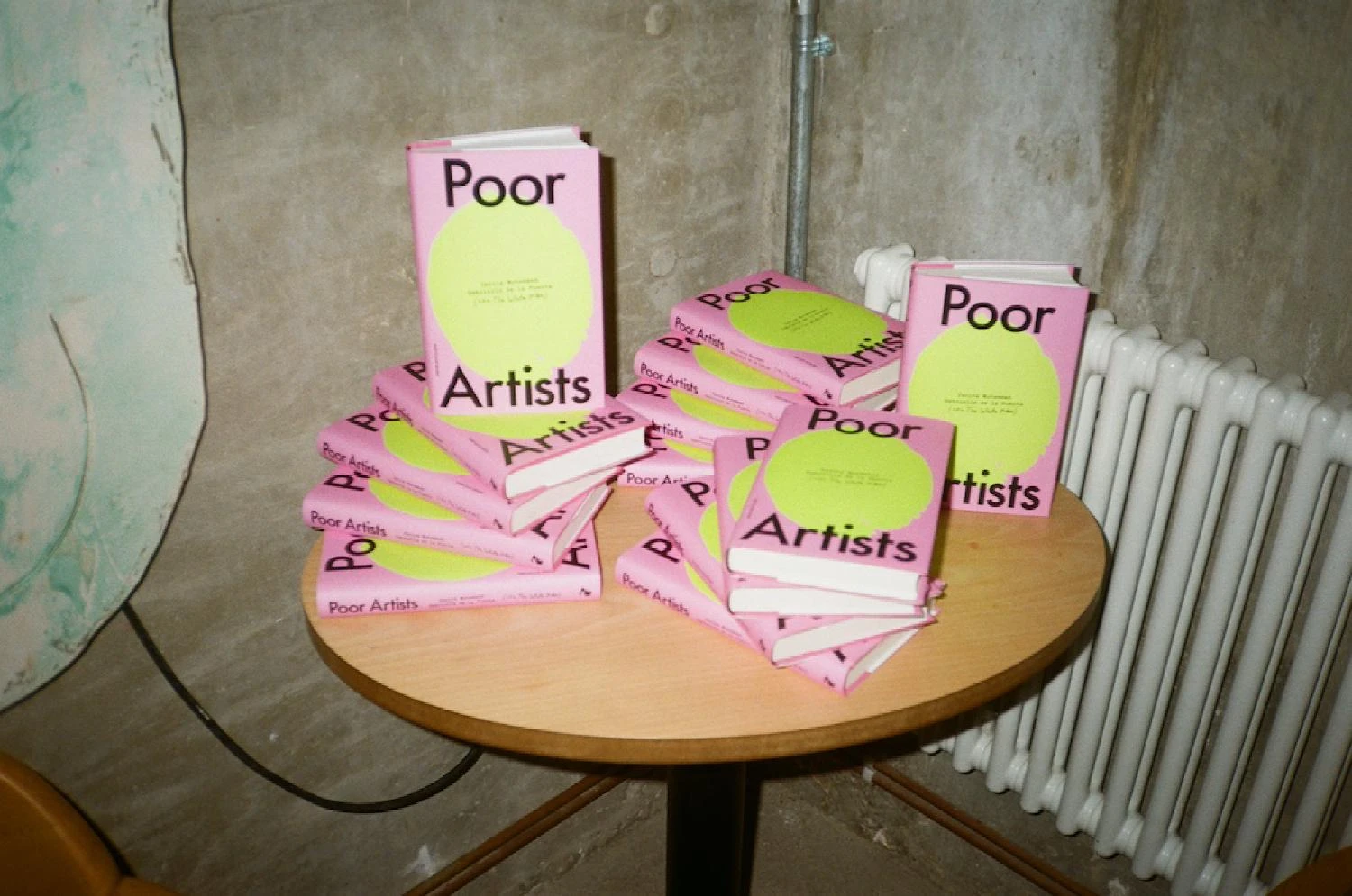Stack of pink copies of Poor Artists by The White Pube arranged on a small round table in a raw concrete space, with a radiator in the background.