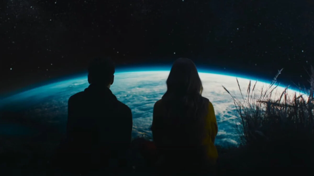 Two silhouetted figures sit side by side, gazing at Earth from space in a still from A Big Bold Beautiful Journey (2025), directed by Kogonada.