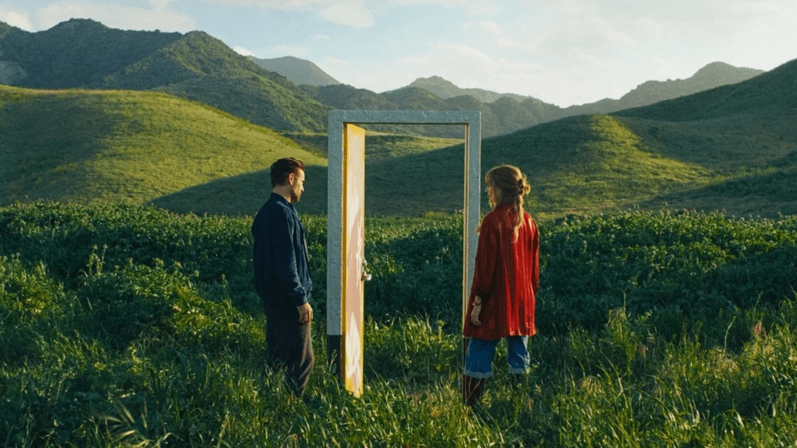 Two characters face each other beside a standing door in a grassy field with rolling hills in a still from A Big Bold Beautiful Journey (2025), directed by Kogonada.
