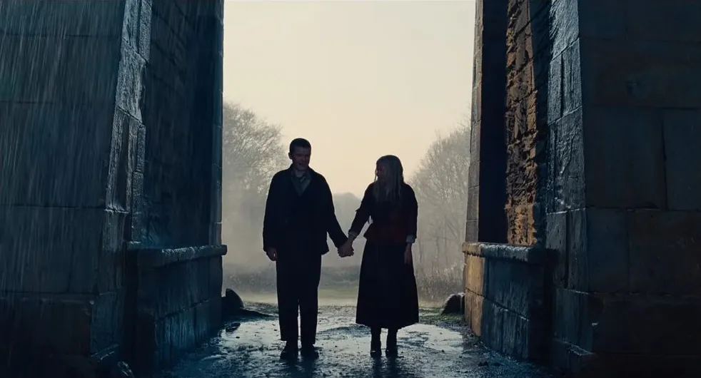 Catherine Earnshaw and Heathcliff walk hand in hand beneath a large stone archway in a cinematic still from Wuthering Heights, silhouetted against soft misty light in a rural landscape. Courtesy of Warner Bros. Pictures