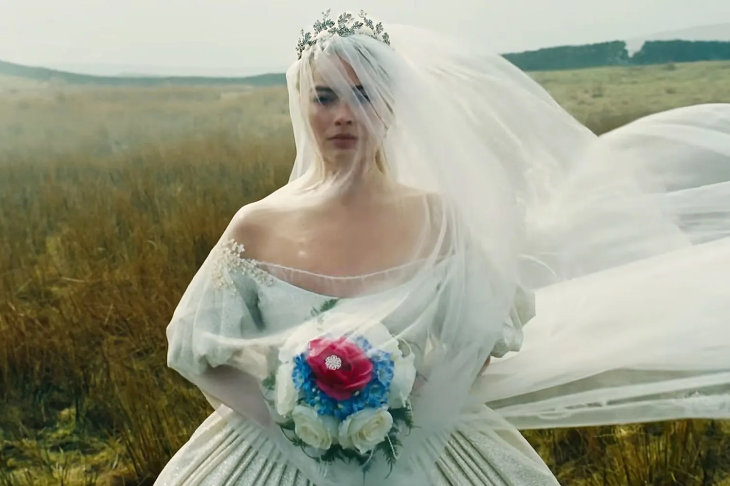 Margot Robbie as Catherine Earnshaw in Wuthering Heights, standing in an open moorland landscape wearing a flowing bridal gown and veil, holding a bouquet as wind moves through the scene in a romantic cinematic still