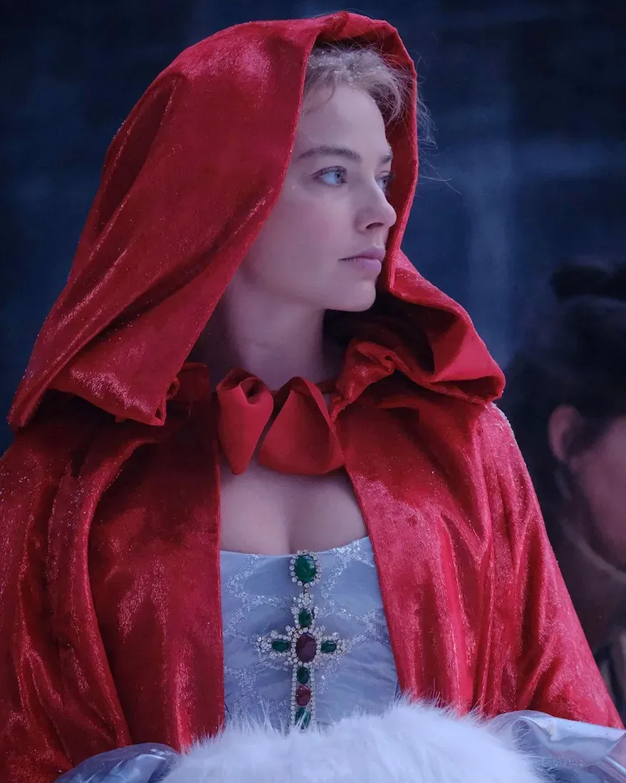 Catherine Earnshaw, portrayed by Margot Robbie, wearing a vivid red hooded cloak over a jeweled period dress, shown in a cinematic close up from Wuthering Heights against a cool toned atmospheric background