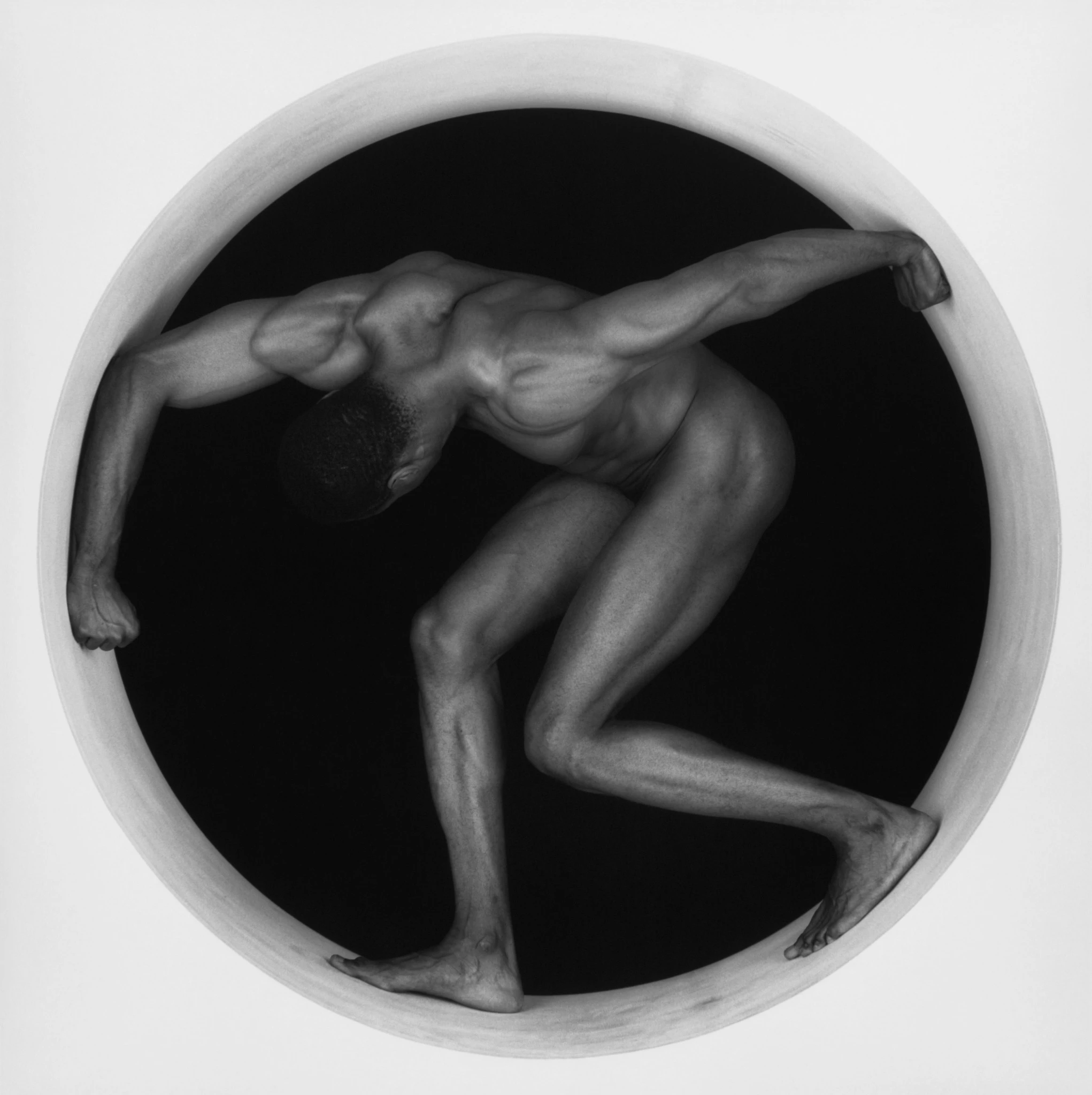Black and white photograph of a muscular nude male figure crouched inside a circular frame, his body forming a dynamic sculptural pose against a dark background. Thomas, 1987 Robert Mapplethorpe Foundation. Used by permission