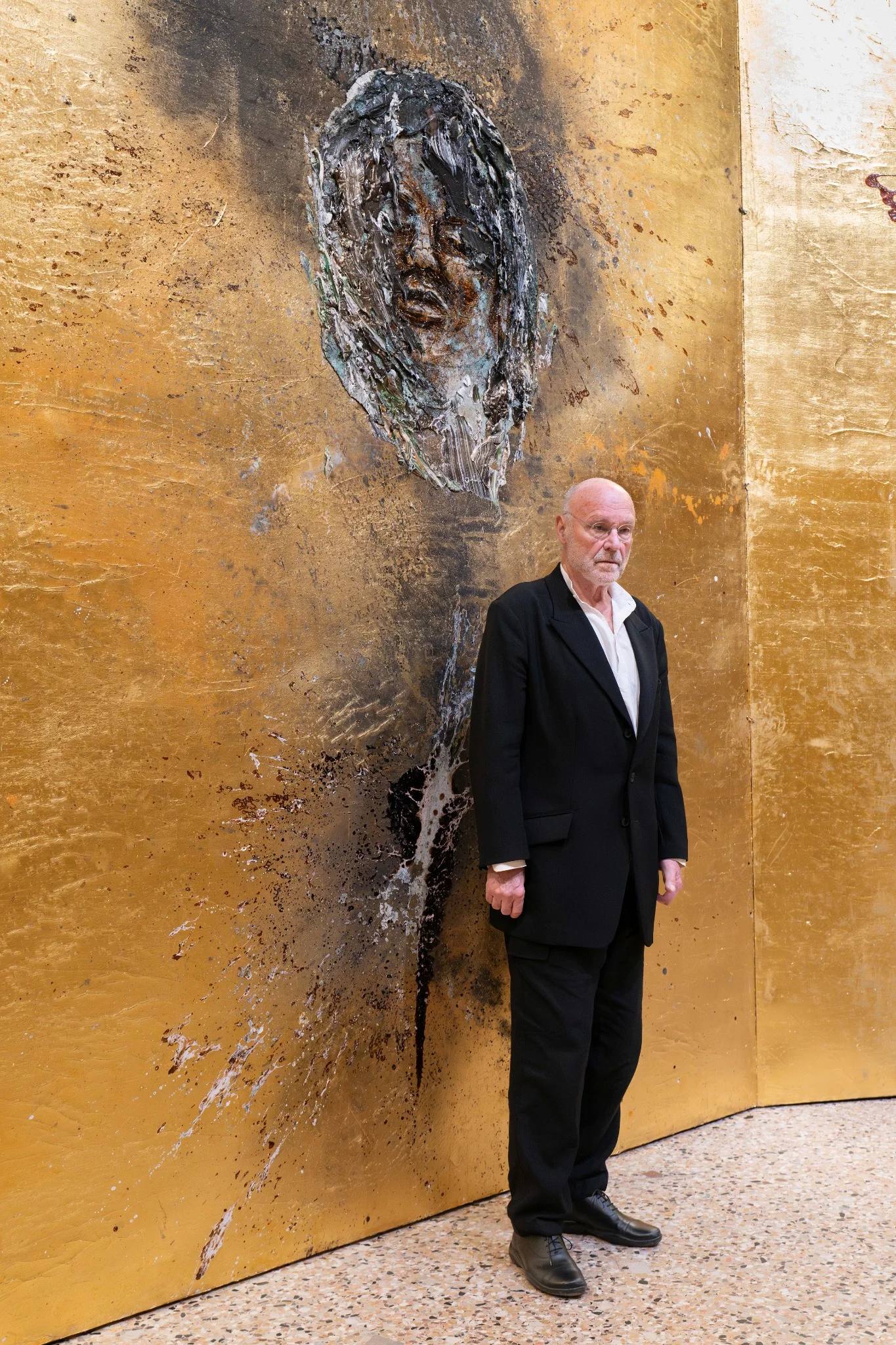 Portrait of Anselm Kiefer standing before a large textured painting with gold surface and sculptural face, photographed at Palazzo Reale, Milan.