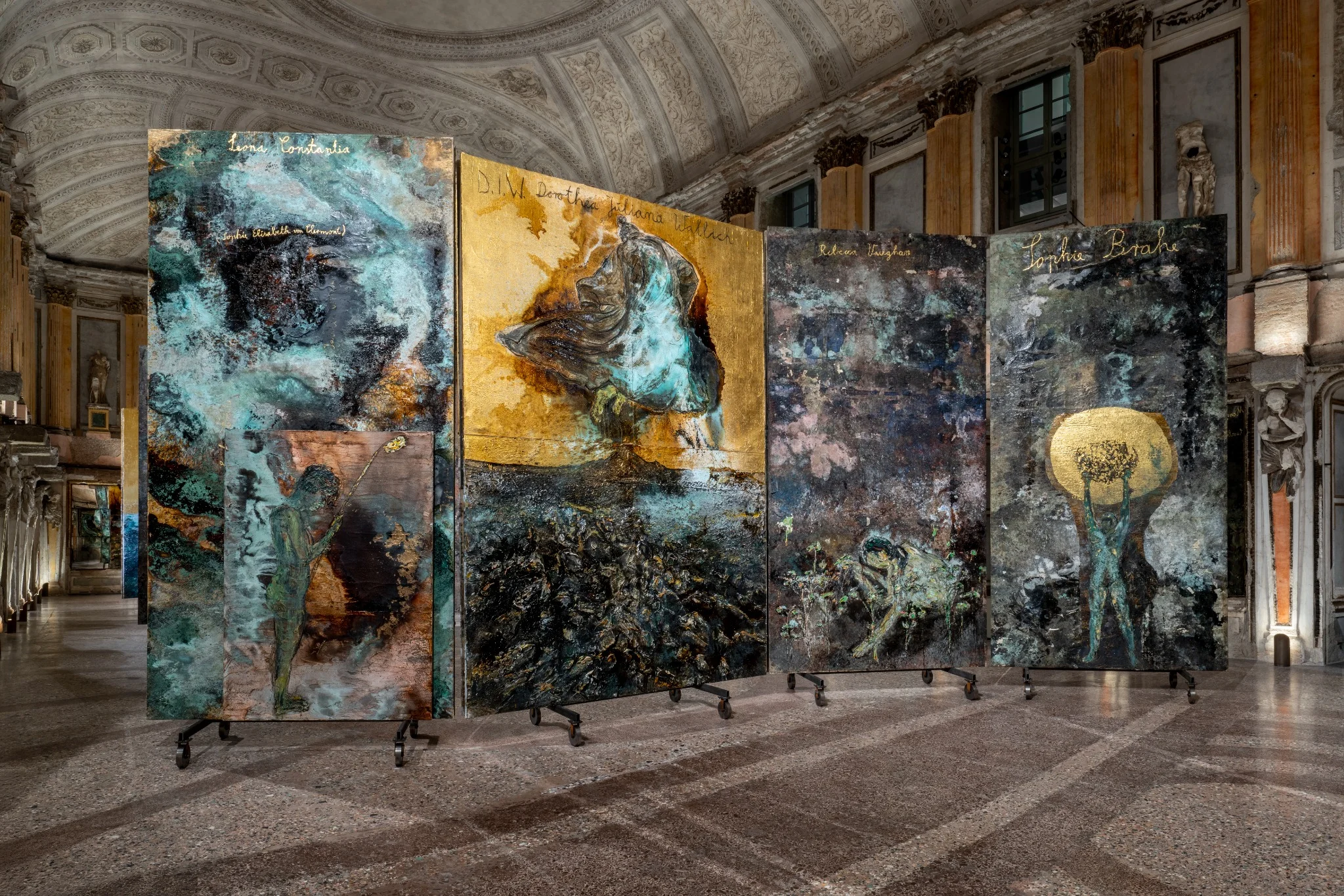 Installation view of Anselm Kiefer’s “The Women Alchemists” at Palazzo Reale, Sala delle Cariatidi, Milan, featuring large-scale textured panels with alchemical and figurative imagery.