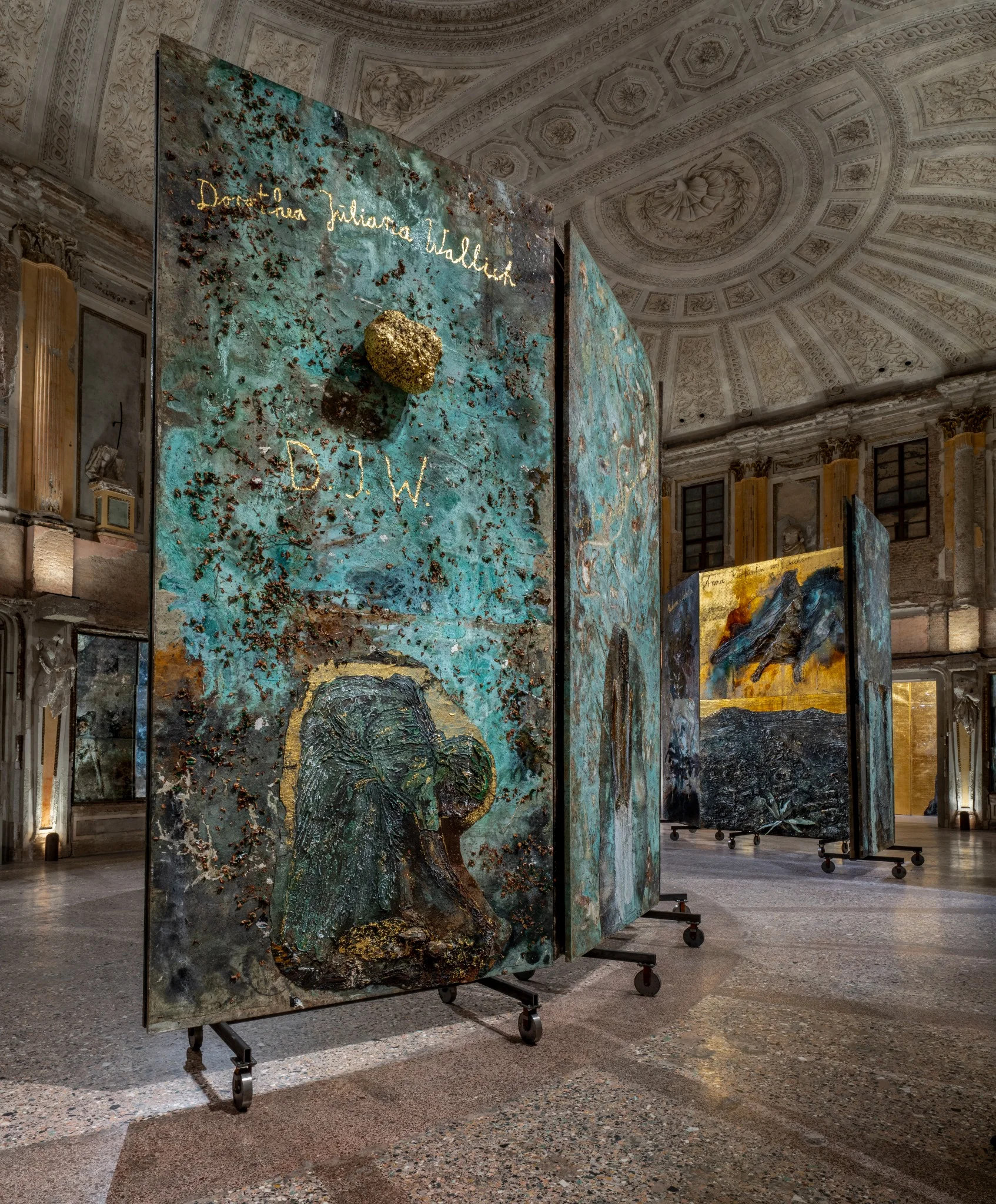 Installation view of Anselm Kiefer’s “The Women Alchemists” at Palazzo Reale, Sala delle Cariatidi, Milan, featuring large-scale textured panels with alchemical and figurative imagery.