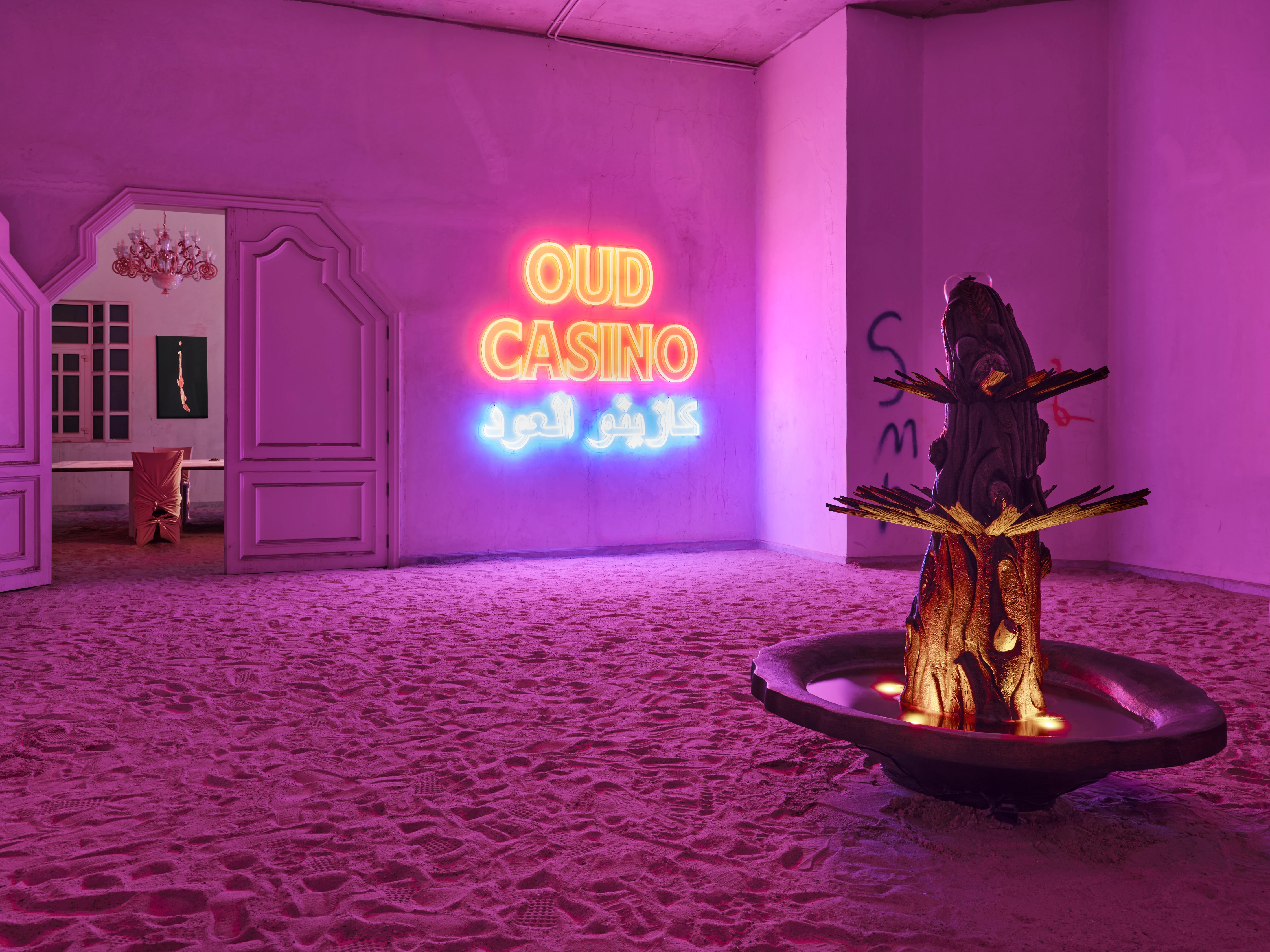Oud Casino neon sign glowing pink over sand-covered gallery floor with sculptural palm tree fountain