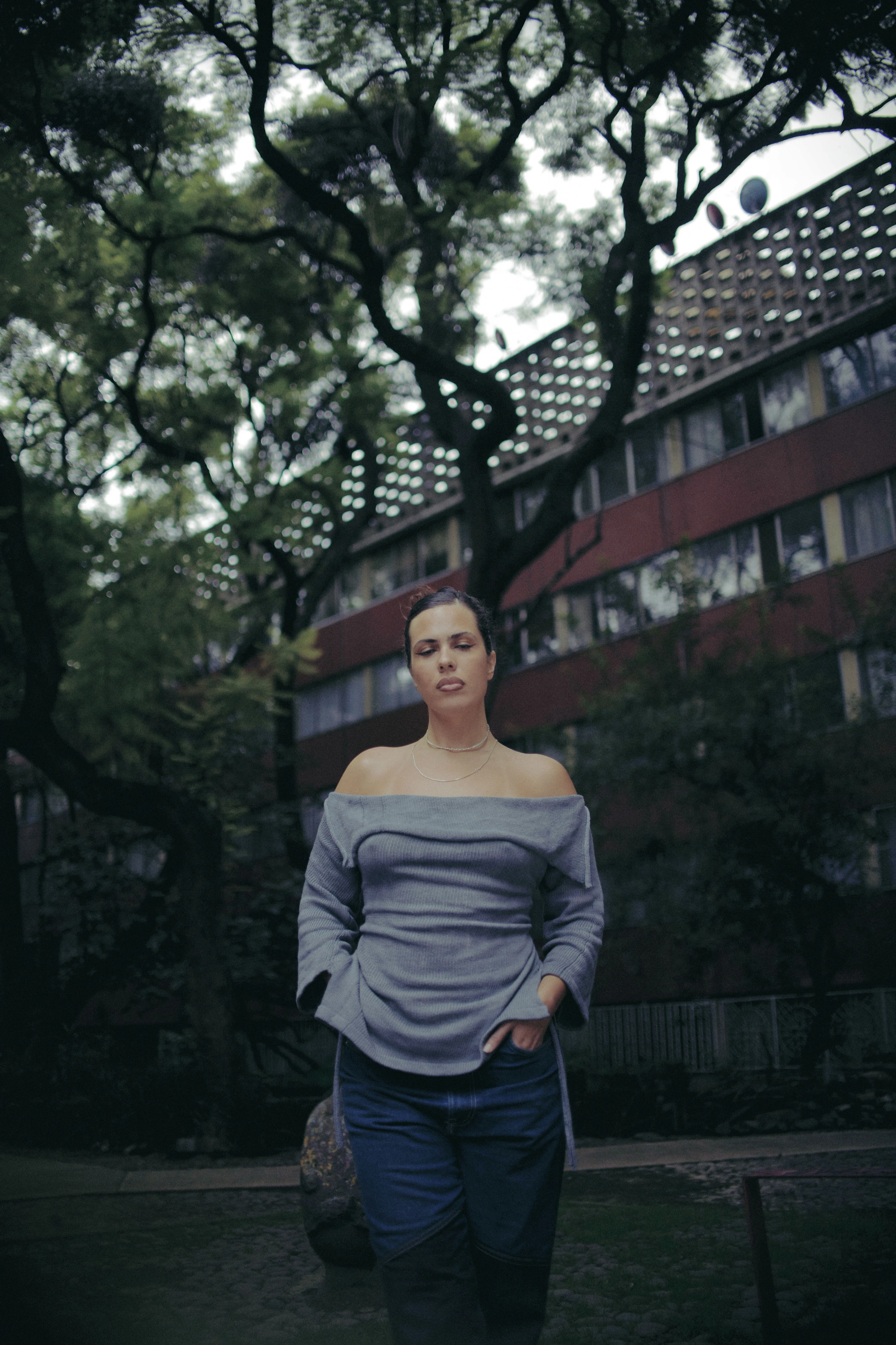 Paloma Contreras Lomas in gray off-shoulder top and jeans standing outdoors beneath lush trees and modernist building