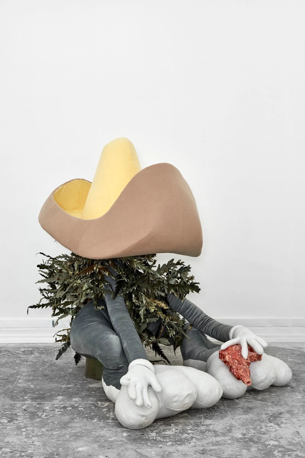 Paloma Contreras Lomas sculpture: seated figure with oversized cowboy hat, fern collar, gray limbs, and white gloved hands holding raw meat