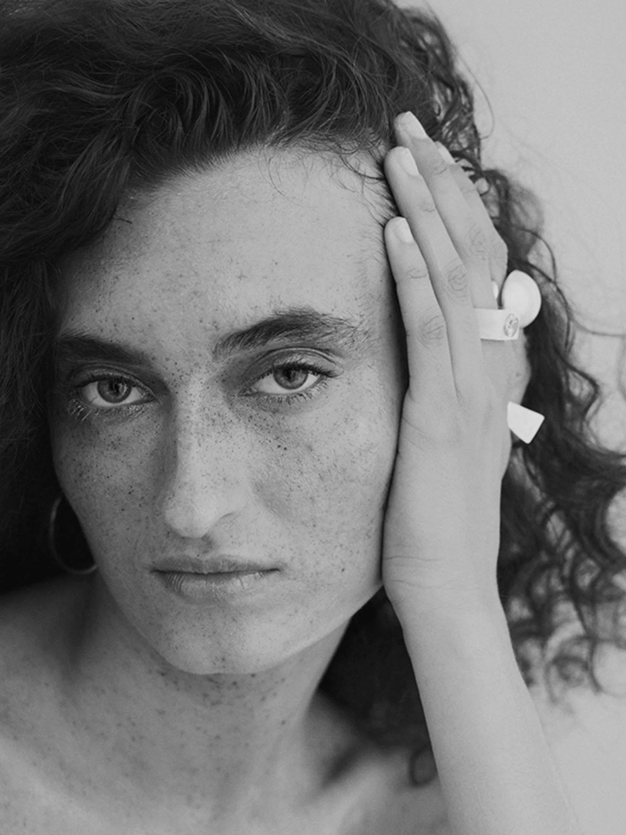 Matterica close-up portrait, woman with freckles and curly hair resting hand on face, wearing sculptural ring