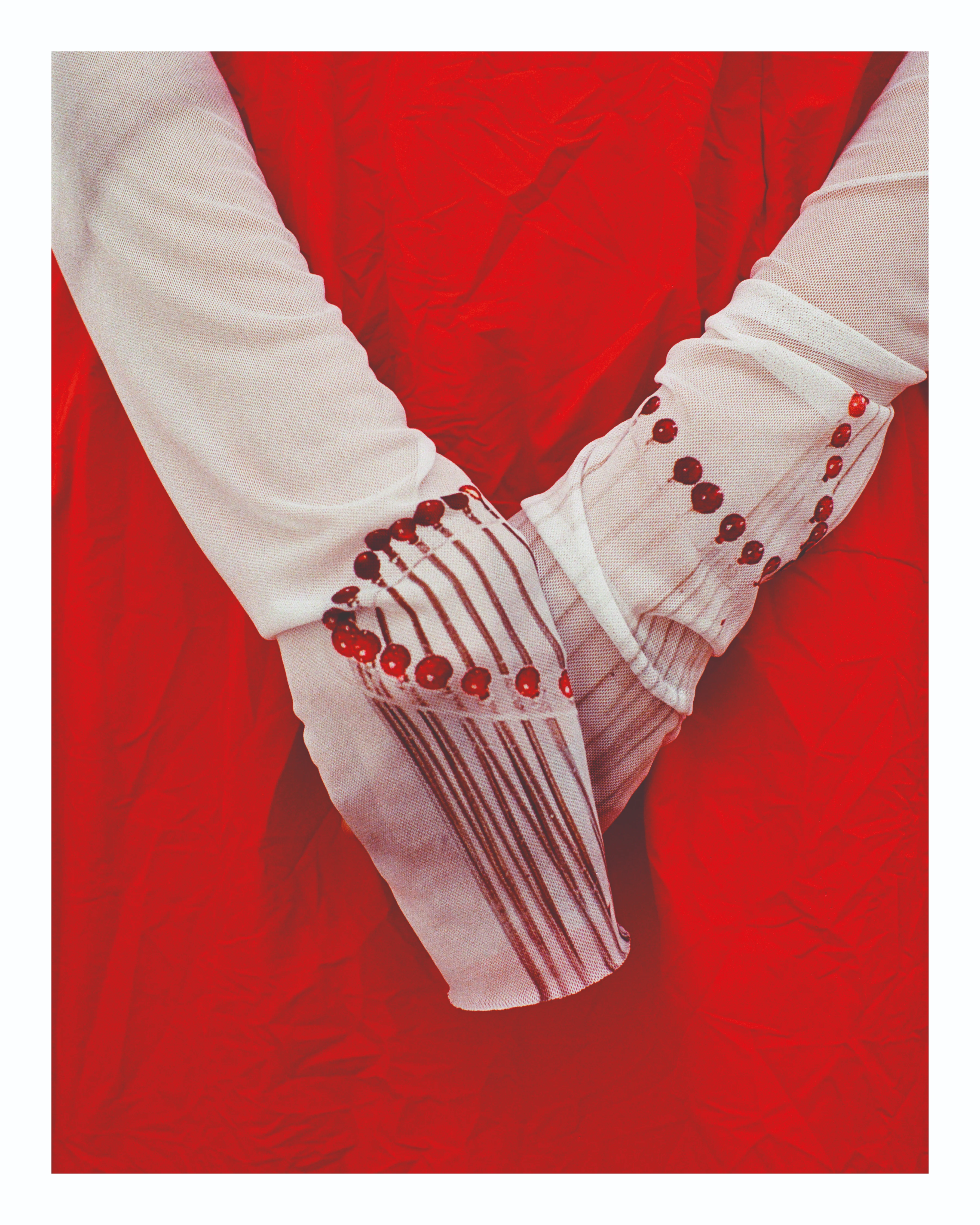 Maria Calderara SS27 Xilema white linen cuffs with red bead embroidery, hands clasped over crimson velvet