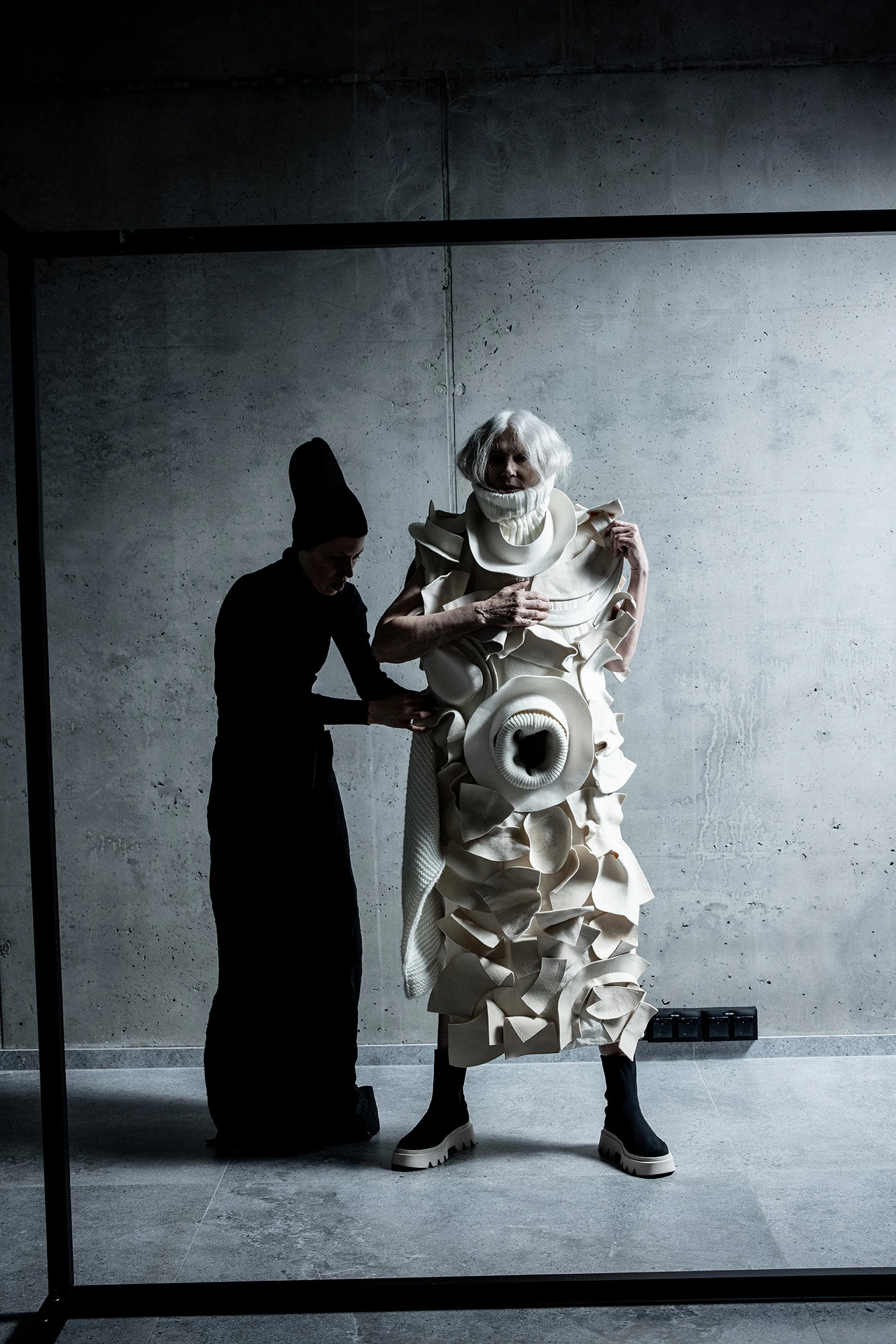 DZHUS ABSOLUTE collection: model in sculptural white ruffled coat assisted by dresser in black, concrete studio setting