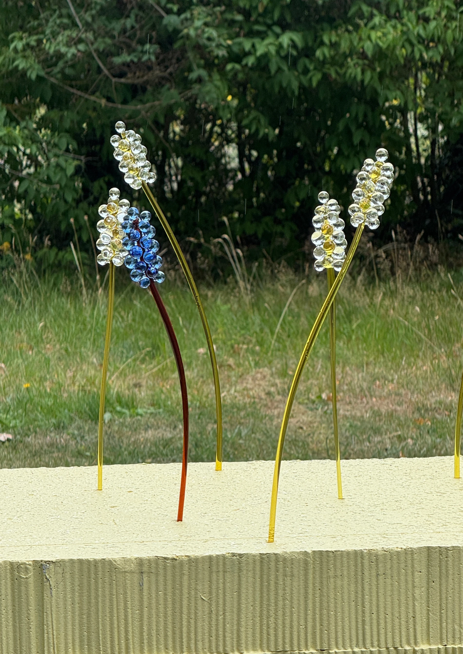 MANU Meadow Collection glass bead flower sculptures on concrete ledge with green meadow backdrop