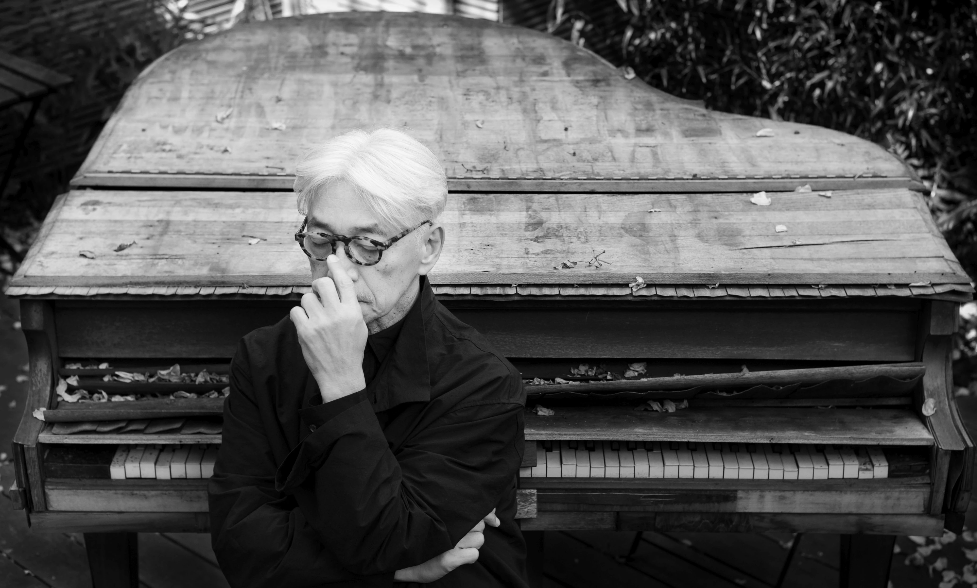 Ryuichi Sakamoto seated pensively before a weathered grand piano, black-and-white portrait by Neo Sora, 2020