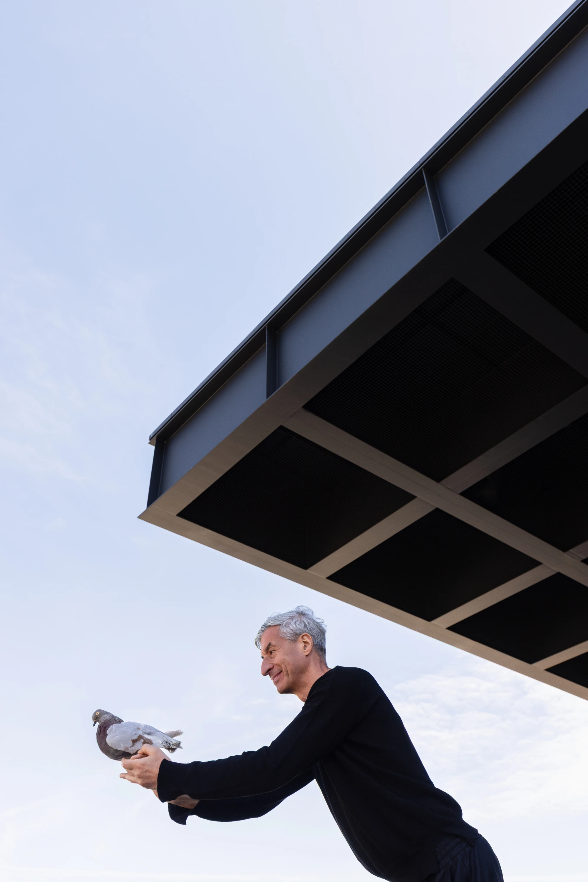 Maurizio Cattelan holding a pigeon outdoors beneath a dark architectural canopy, Neue Nationalgalerie 2026