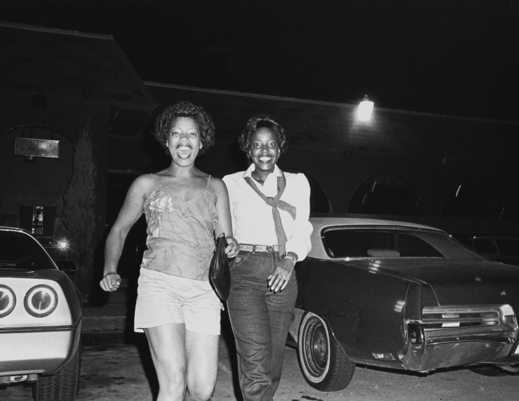Two women laughing together in a parking lot at night, from Carrie Mae Weems' Welcome Home series, 1978–84