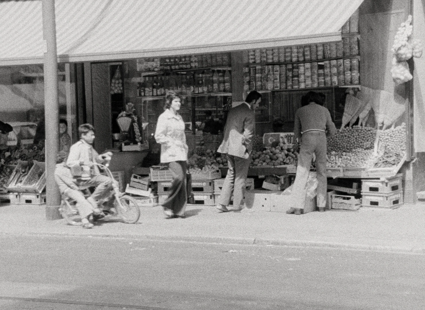 Black-and-white street scene outside a fruit and vegetable shop, 1975 film still by Mehrangis Montazami-Dabui