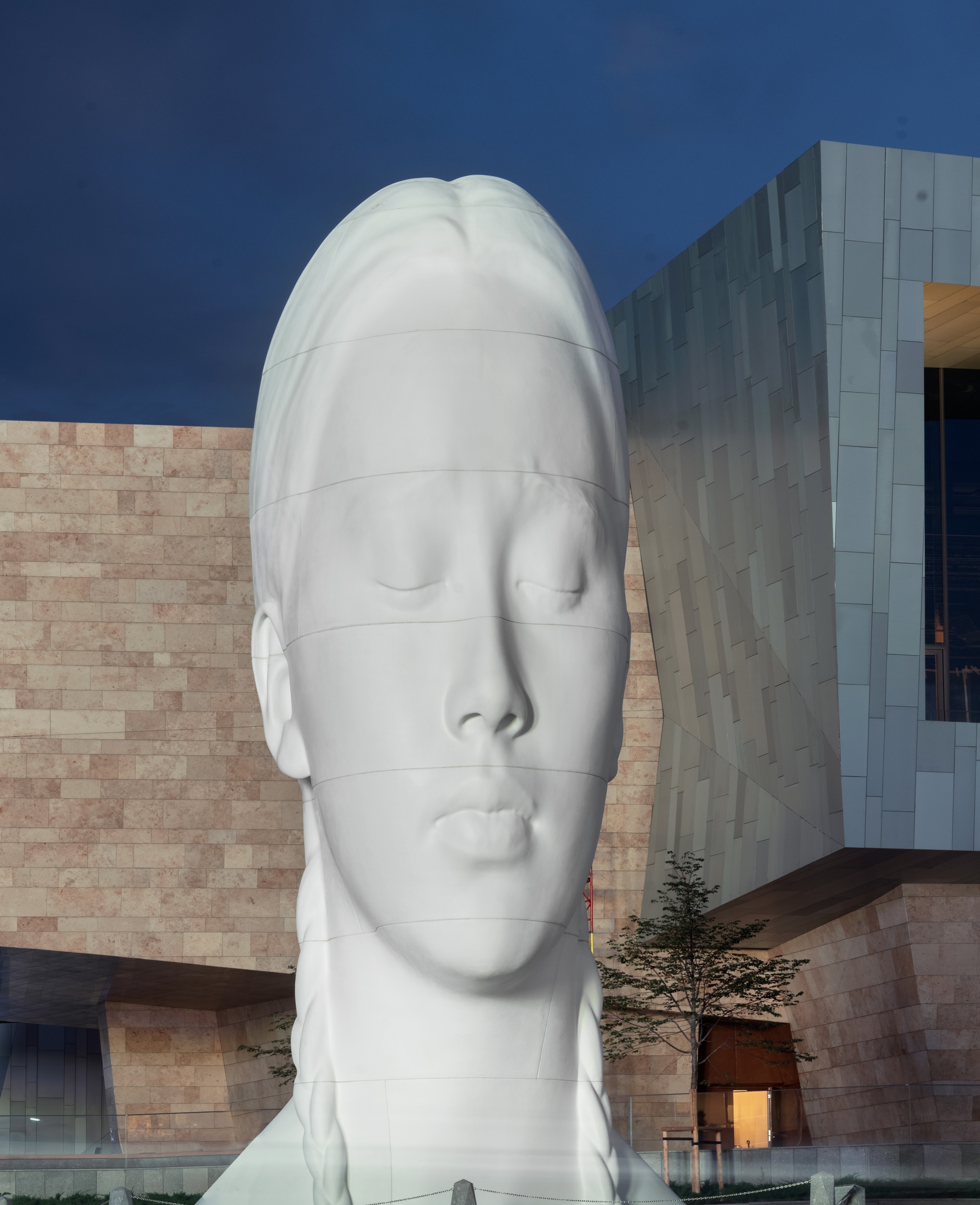 Large white sculptural head of a serene face with closed eyes and horizontal segmented lines, standing outdoors in front of a modern angular museum building with stone and metal cladding, photographed at dusk against a deep blue sky. Jaume Plensa Almaty Kazakhstan
