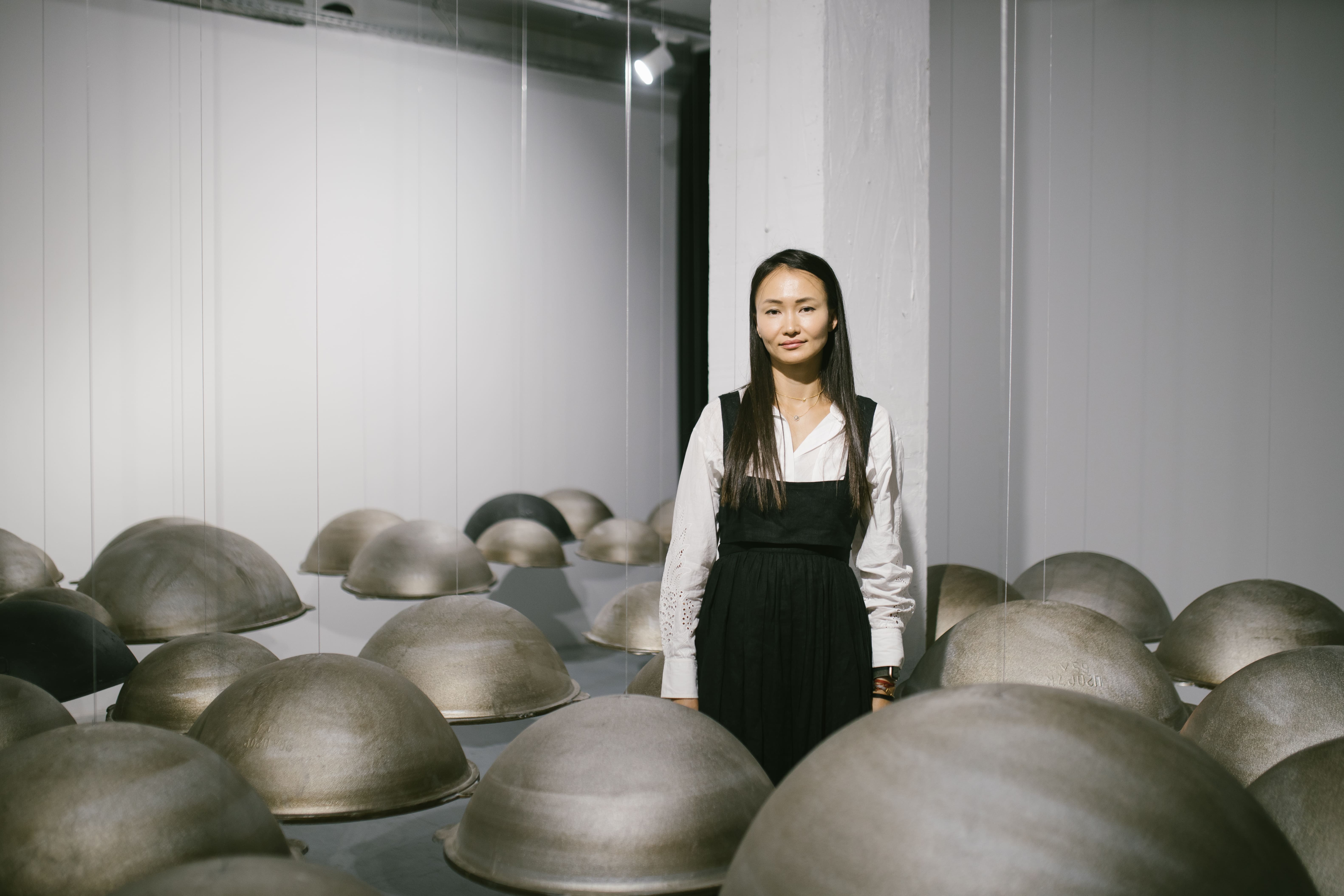 A woman Meruyert Kaliyeva with long dark hair wearing a white blouse and black pinafore dress stands in an art gallery installation surrounded by numerous large dome-shaped metal sculptures suspended from thin wires at varying heights, against white walls. Almaty Kazakhstan