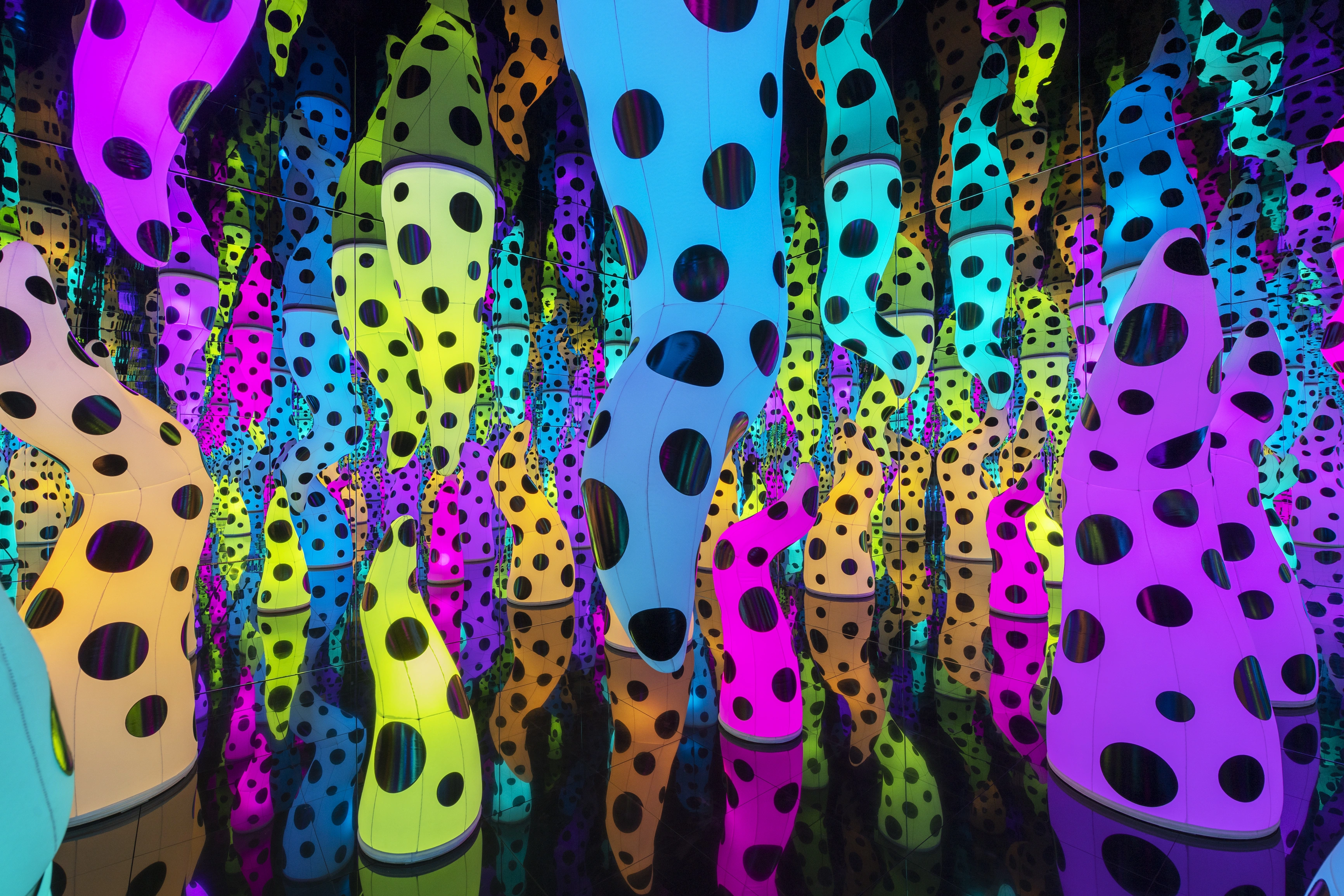 Immersive art installation by Yayoi Kusama, featuring a dense forest of tall, glowing inflatable organic forms in vivid pink, blue, yellow, orange, and purple, each covered in large black polka dots, reflected infinitely in a mirrored room.