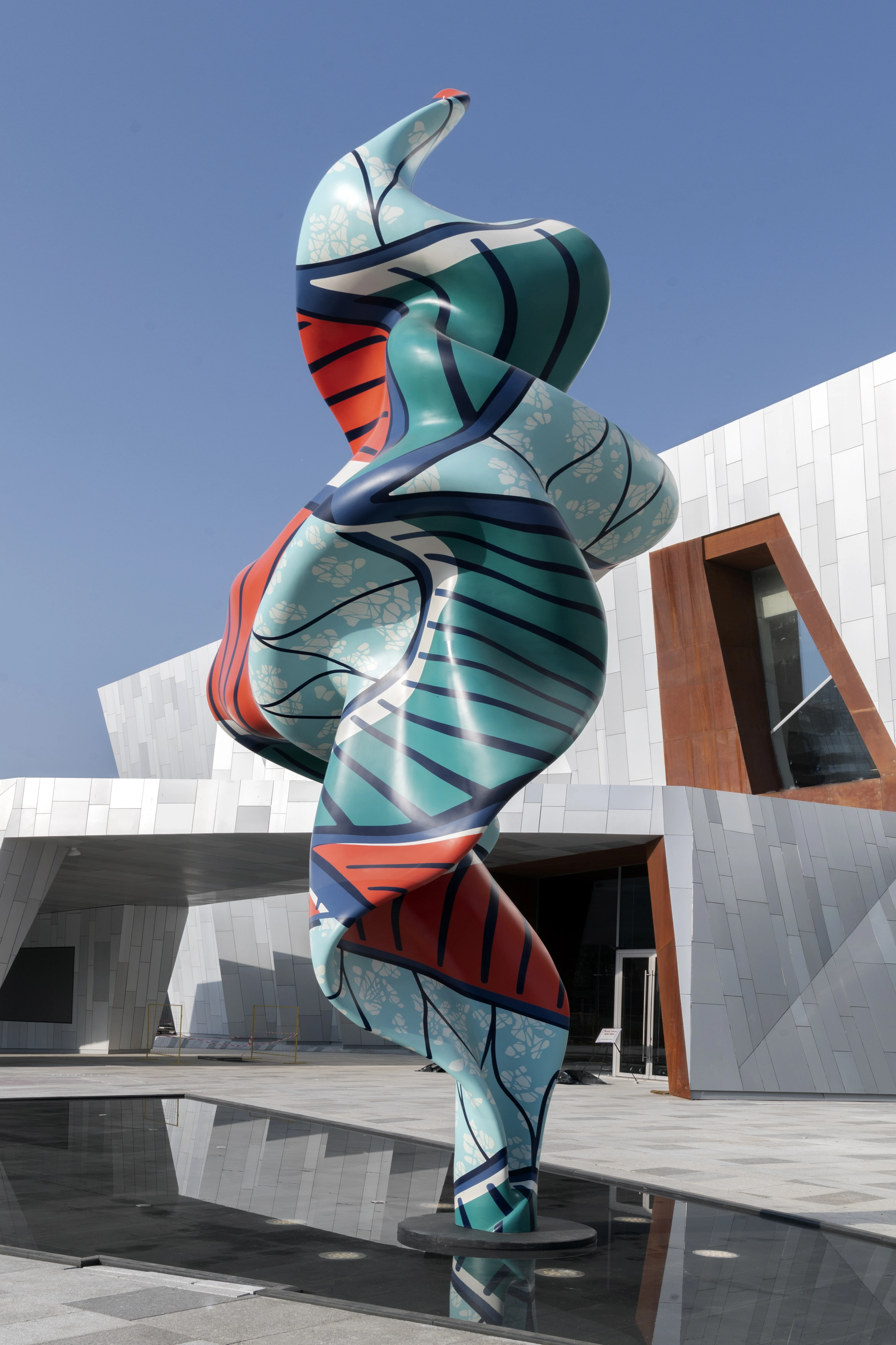 Tall twisting sculpture with a smooth, biomorphic form painted in teal, orange, and navy patterned stripes and floral motifs, standing on a reflective black base in front of a modern white and rust-colored angular museum building under a clear blue sky. Yinka Shonibare