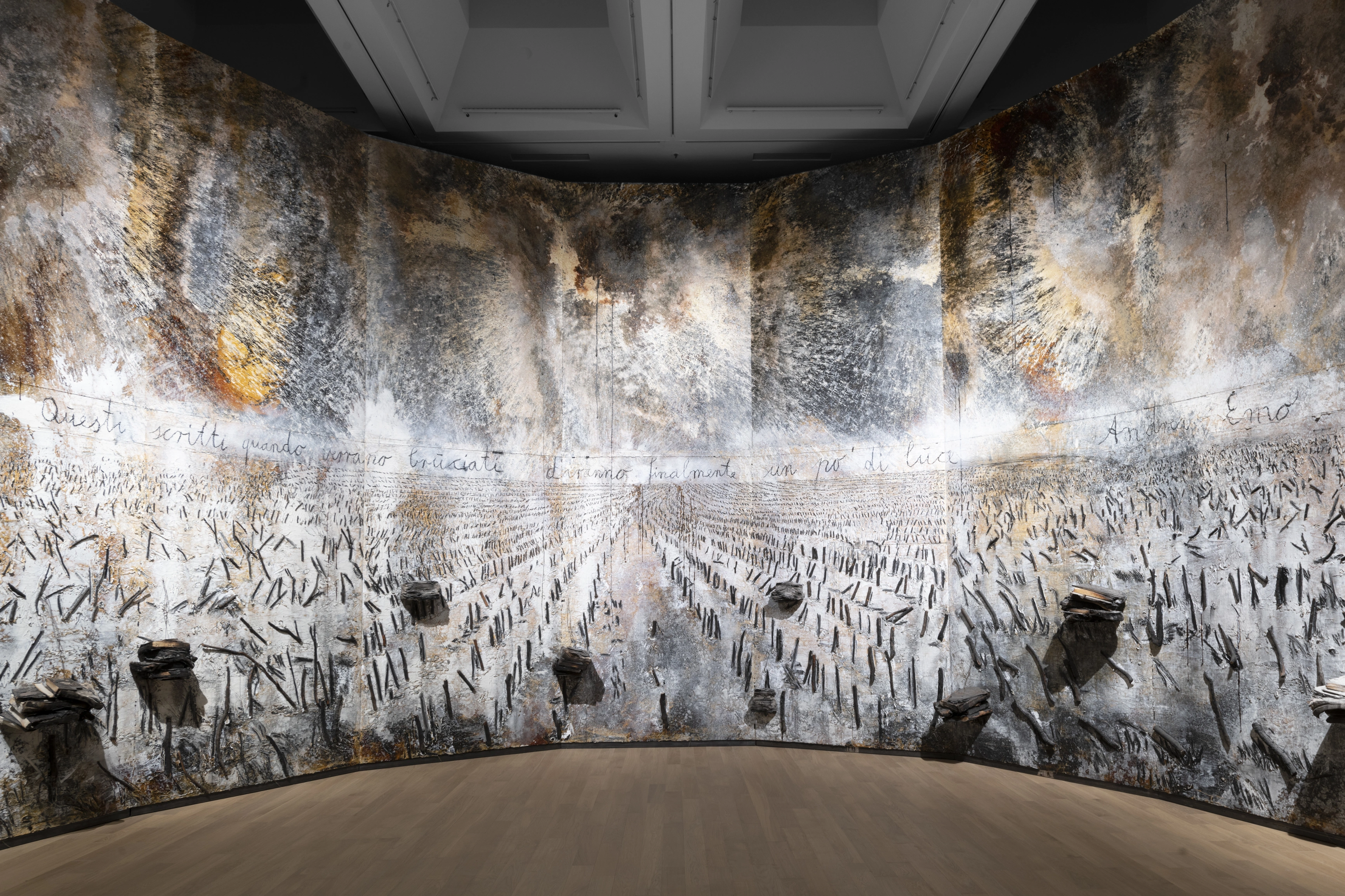Anselm Kiefer, Questi scritti, quando verranno bruciati, daranno Finalmente un po' di luce (2020–2021), Almaty Museum of Arts, Kazakhstan. Image courtesy of the Almaty Museum of Arts, Immersive gallery installation with curved, floor-to-ceiling panels covered in textured burnt and weathered paint in gray, white, and rust tones, resembling a scorched field with rows of charred stubble and stacks of burnt books mounted on the wall, with a handwritten Italian inscription running across the panels signed