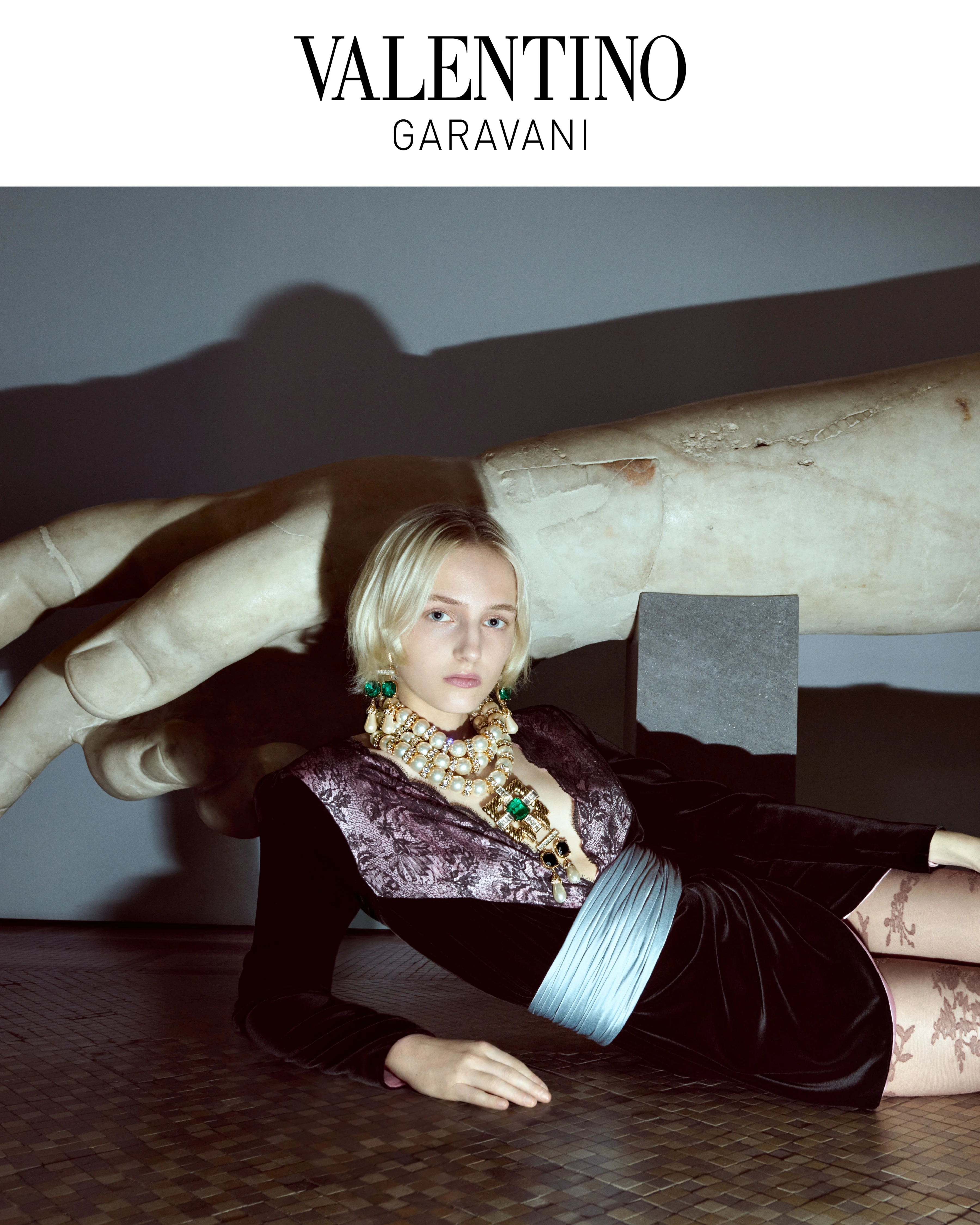 Valentino Garavani FW26 campaign model in velvet dress with gold gemstone necklace beside monumental marble hand sculpture