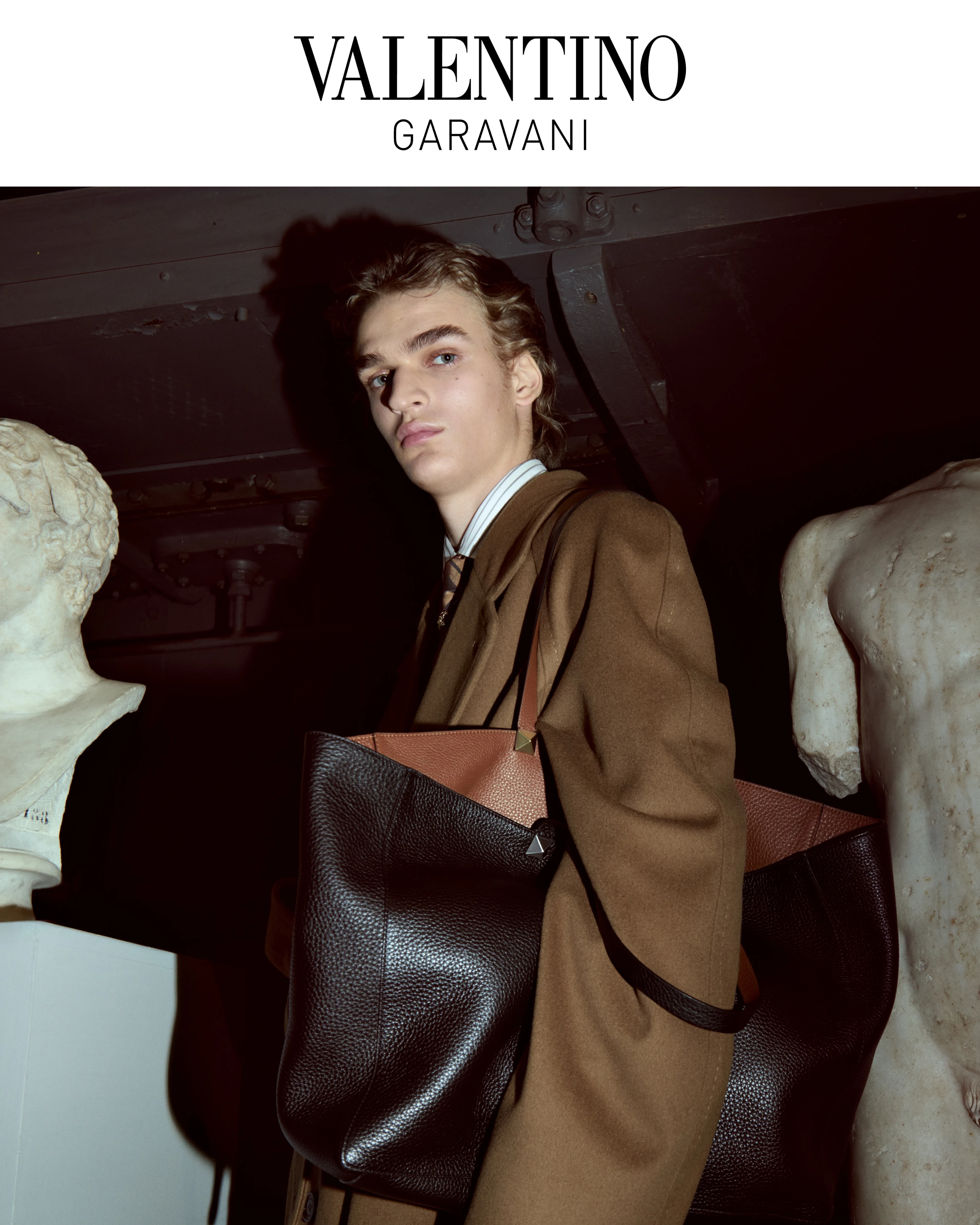 Valentino Garavani FW26 campaign model in brown wool coat carrying oversized dark leather tote bag among classical sculptures