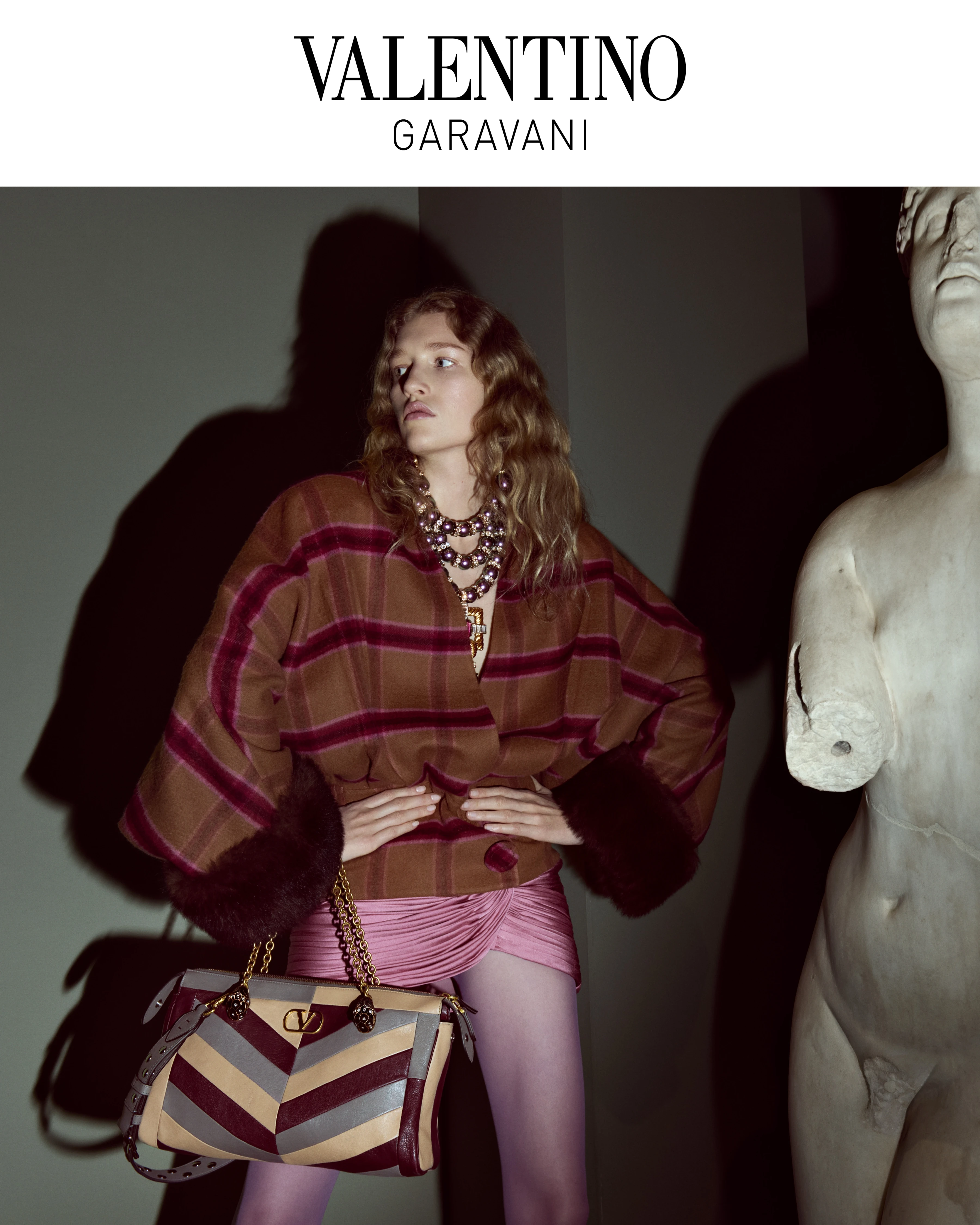 Valentino Garavani FW26 model in plaid cape, pink ruched skirt, layered necklaces, holding striped VLogo chain bag beside classical sculpture