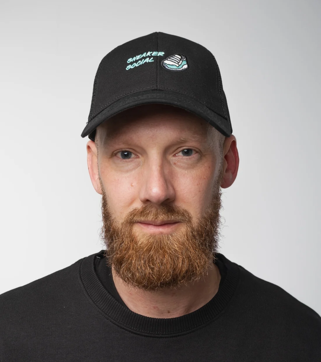 A bearded man faces the camera with a neutral expression, with a black cap atop his head featuring words and an image in blue.