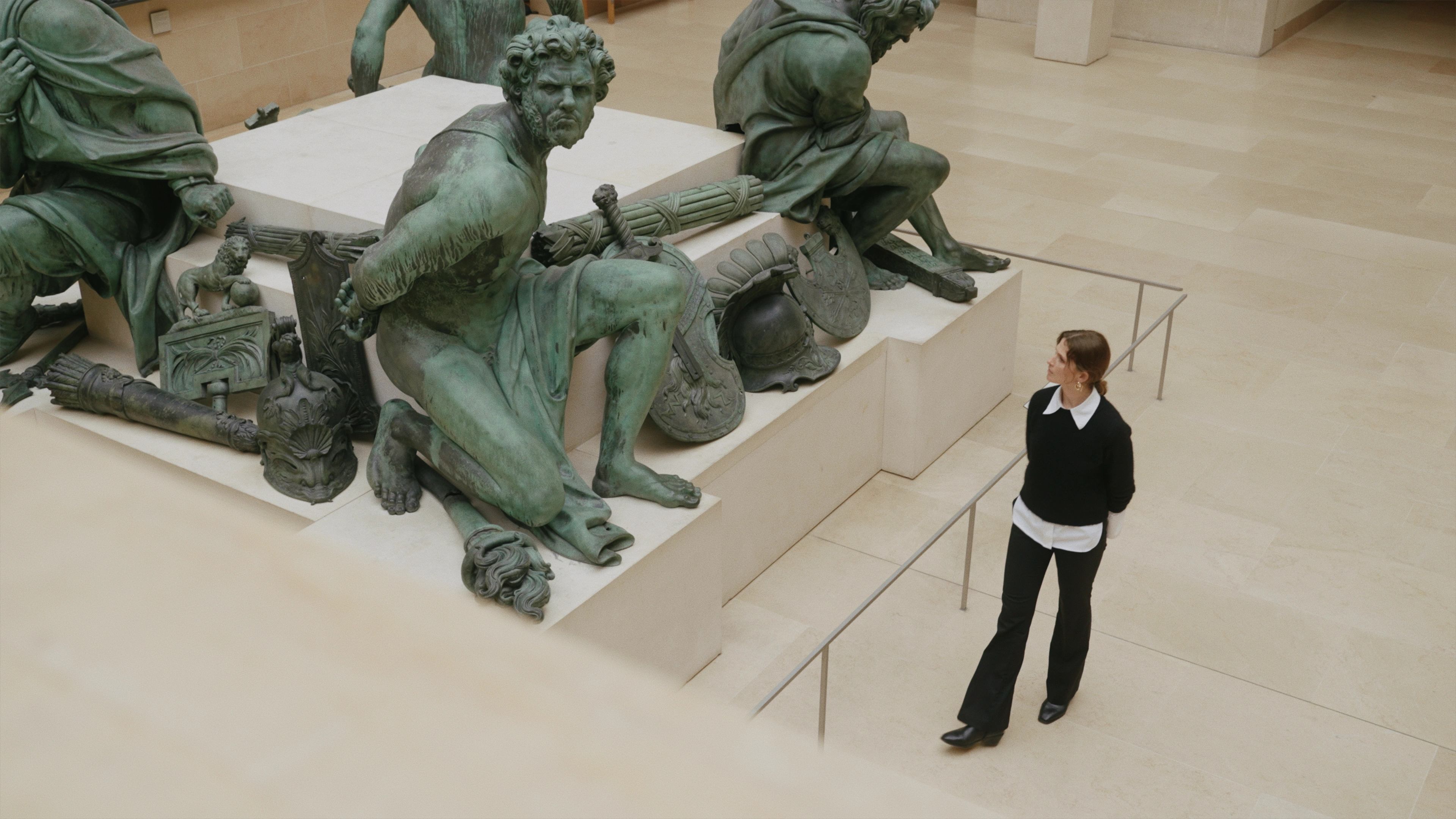 Aerial view of a museum gallery showing a woman in a black sweater and trousers looking up at a large bronze sculptural group of classical warrior figures with weapons, shields, and helmets, displayed on a pale stone pedestal.