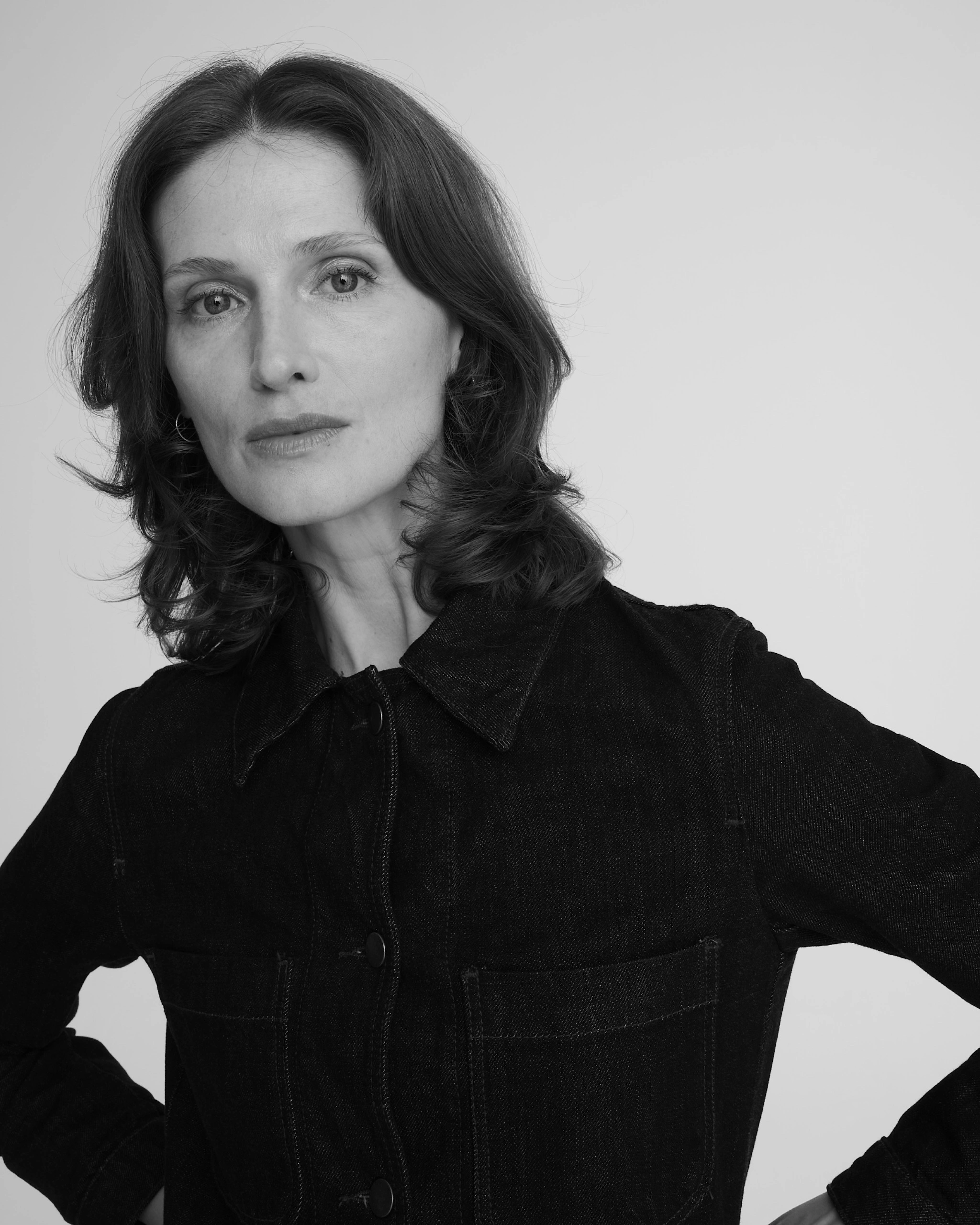 Black-and-white studio portrait of a woman with wavy shoulder-length dark hair, wearing a black denim jacket, standing with hands on her hips, gazing directly at the camera against a plain light background.Marzena Jarczak