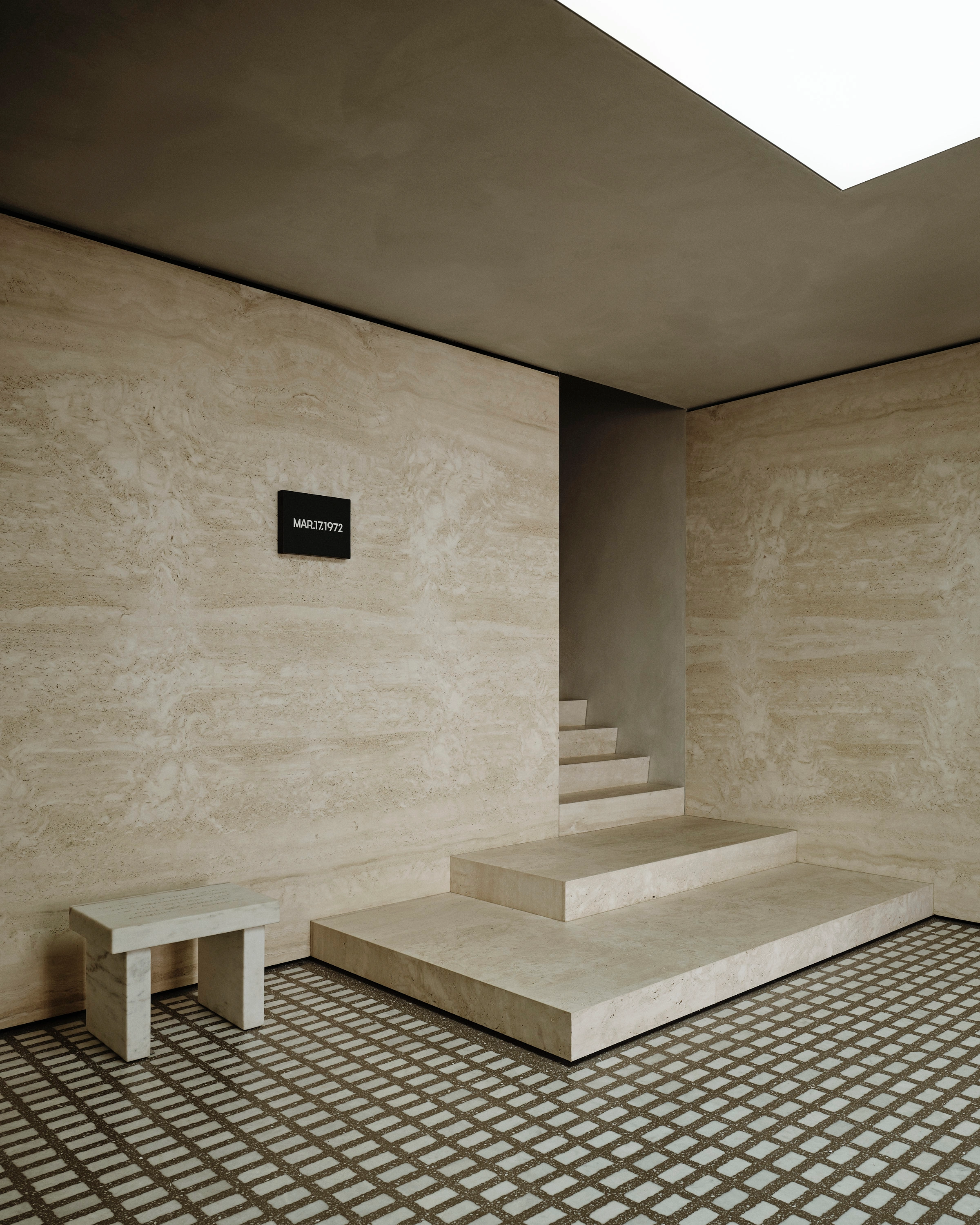 Minimalist travertine-clad interior with a stepped stone platform leading to a narrow staircase, a small black plaque reading 
