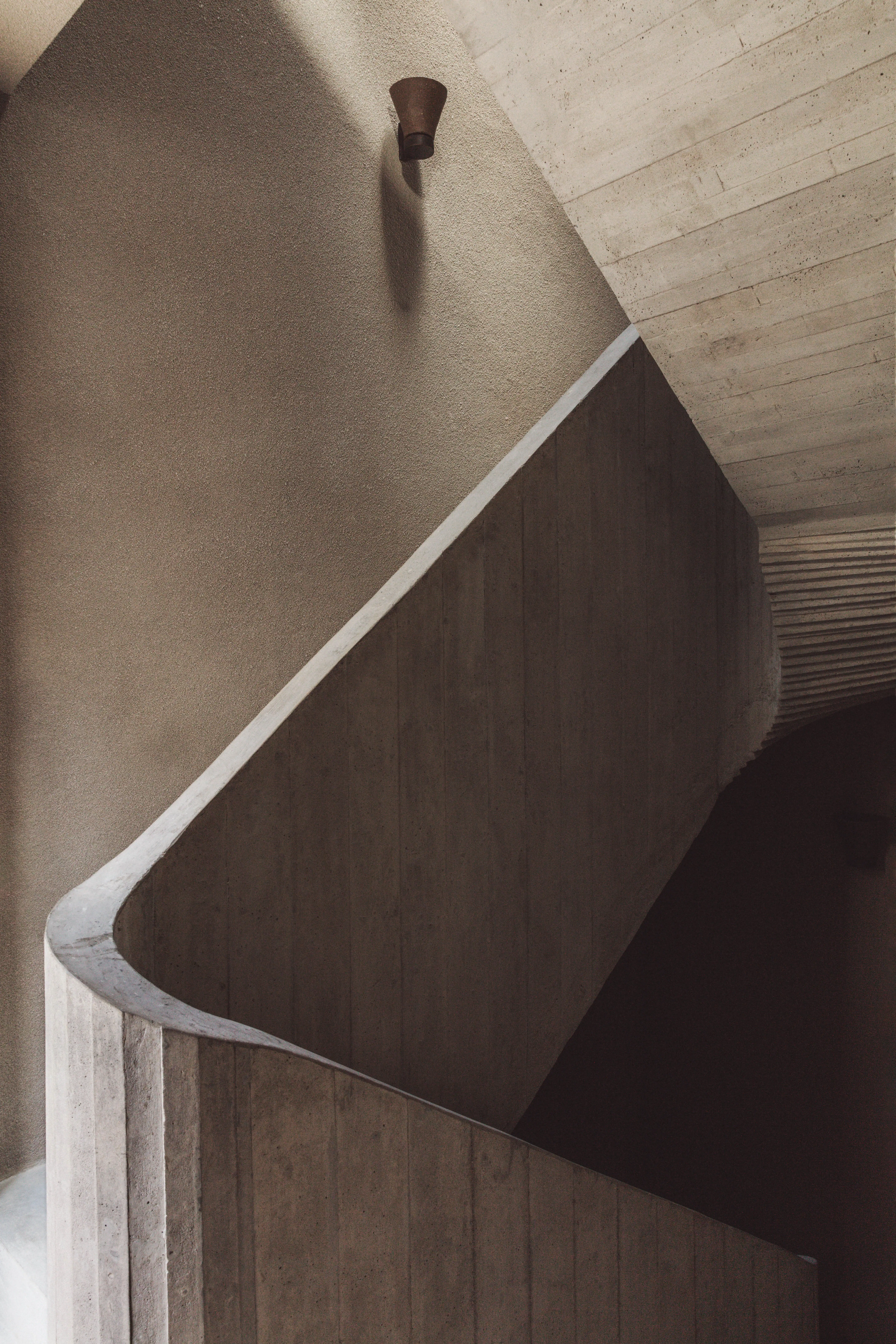 Close-up architectural view of a sculptural concrete staircase with a curved white railing, board-formed textured walls, a ribbed ceiling, and a small bronze wall-mounted sconce casting a shadow. Nicolas Schuybroek